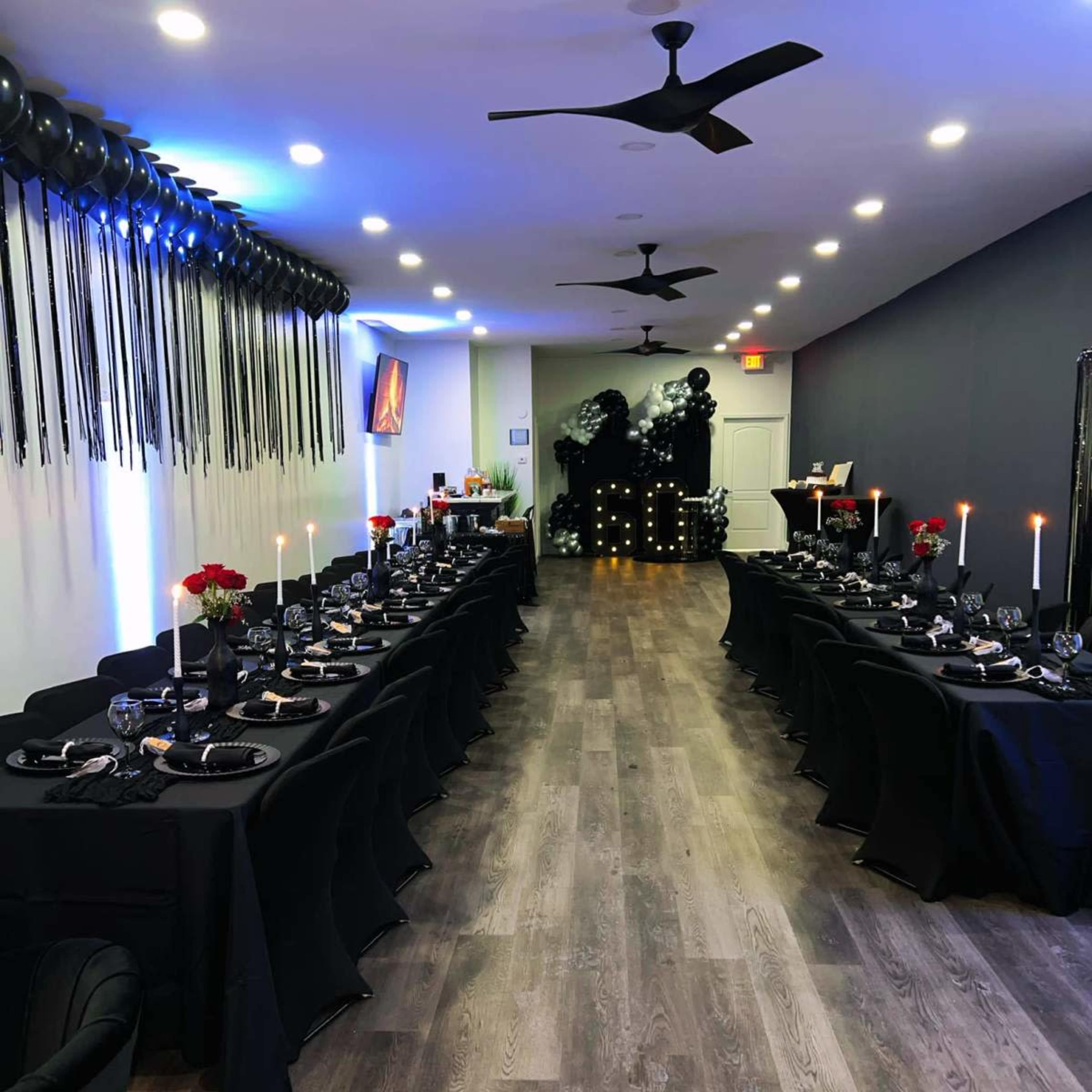 A long, elegantly set dining table with black tablecloths is arranged in a dimly lit room, featuring decorative candles, black balloons, and a wall adorned with dark accents.