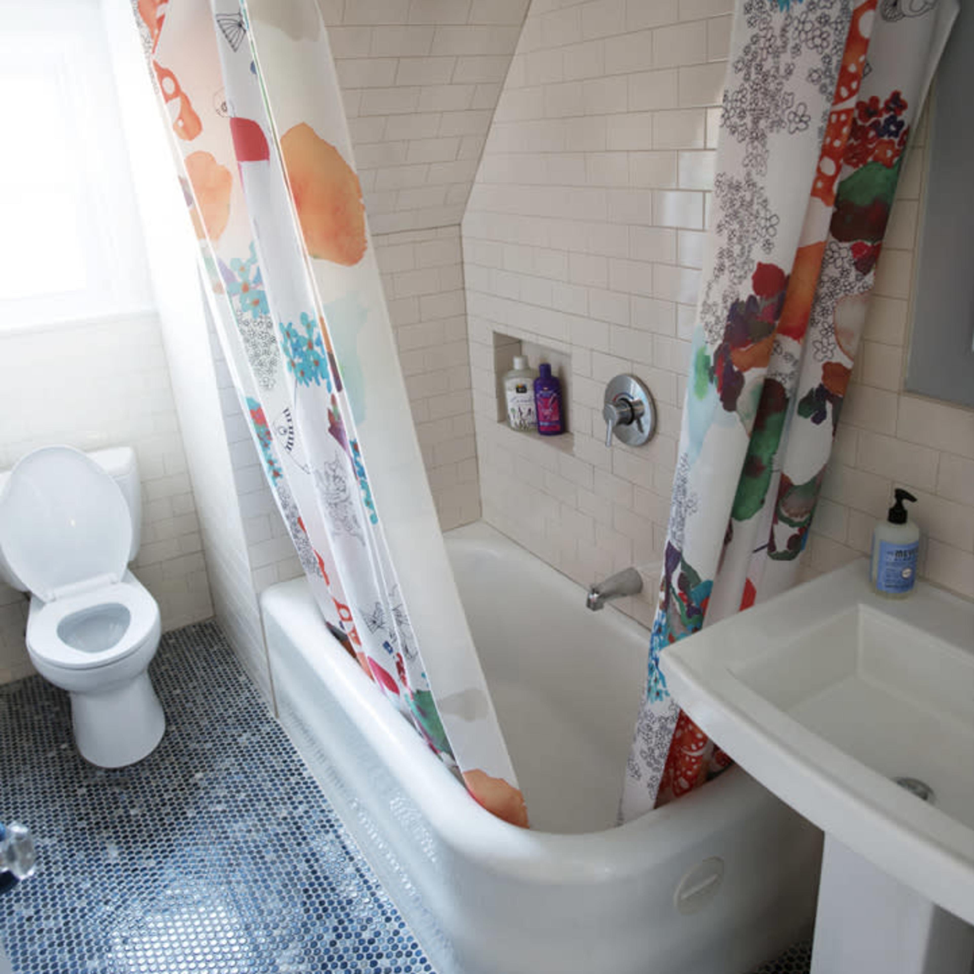 A bathroom features a bathtub with a floral shower curtain, a toilet, and a sink, all set against a tiled wall and mosaic floor.