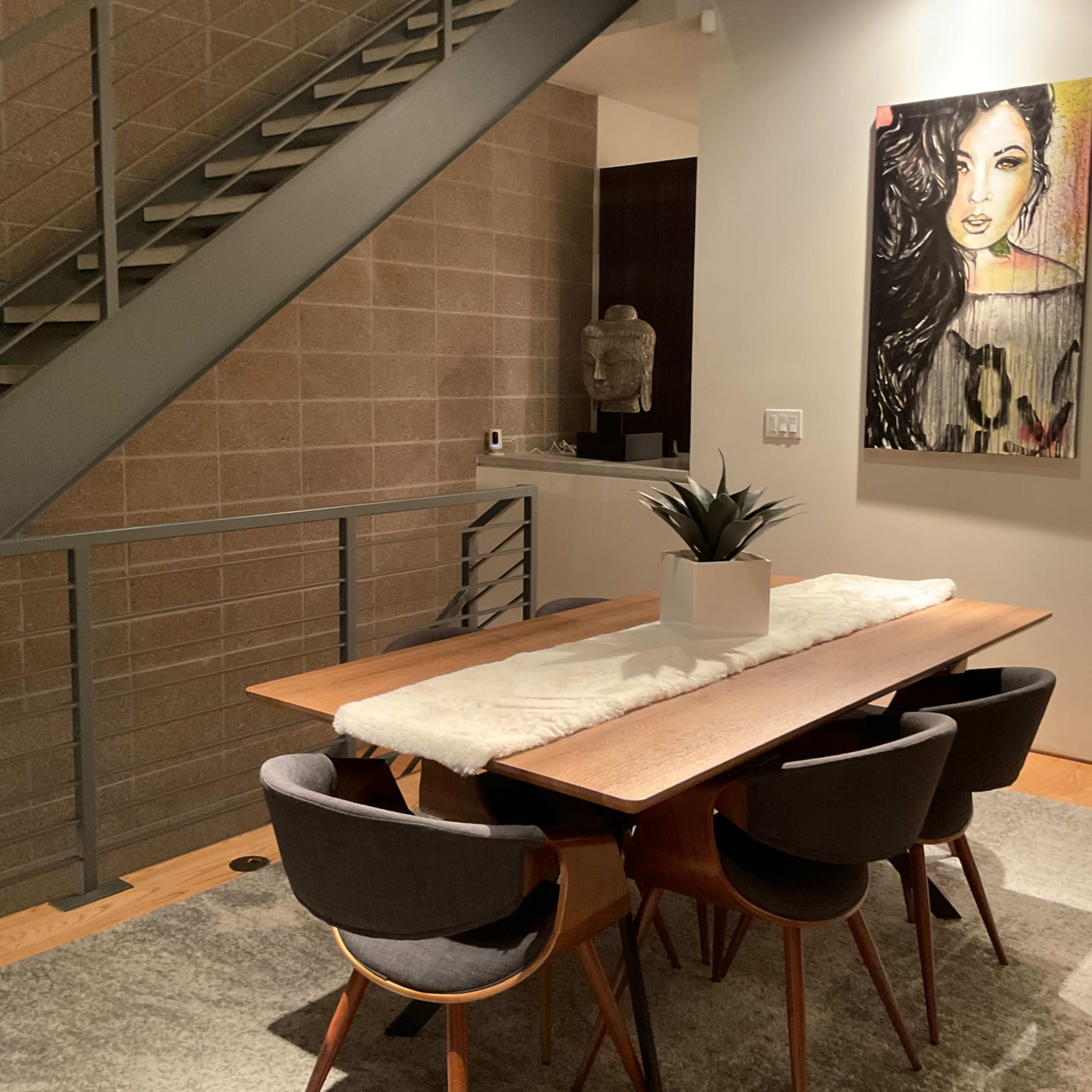 A dining area features a wooden table with upholstered chairs, a decorative plant in the center, a large artwork on the wall, and staircase access in the background.
