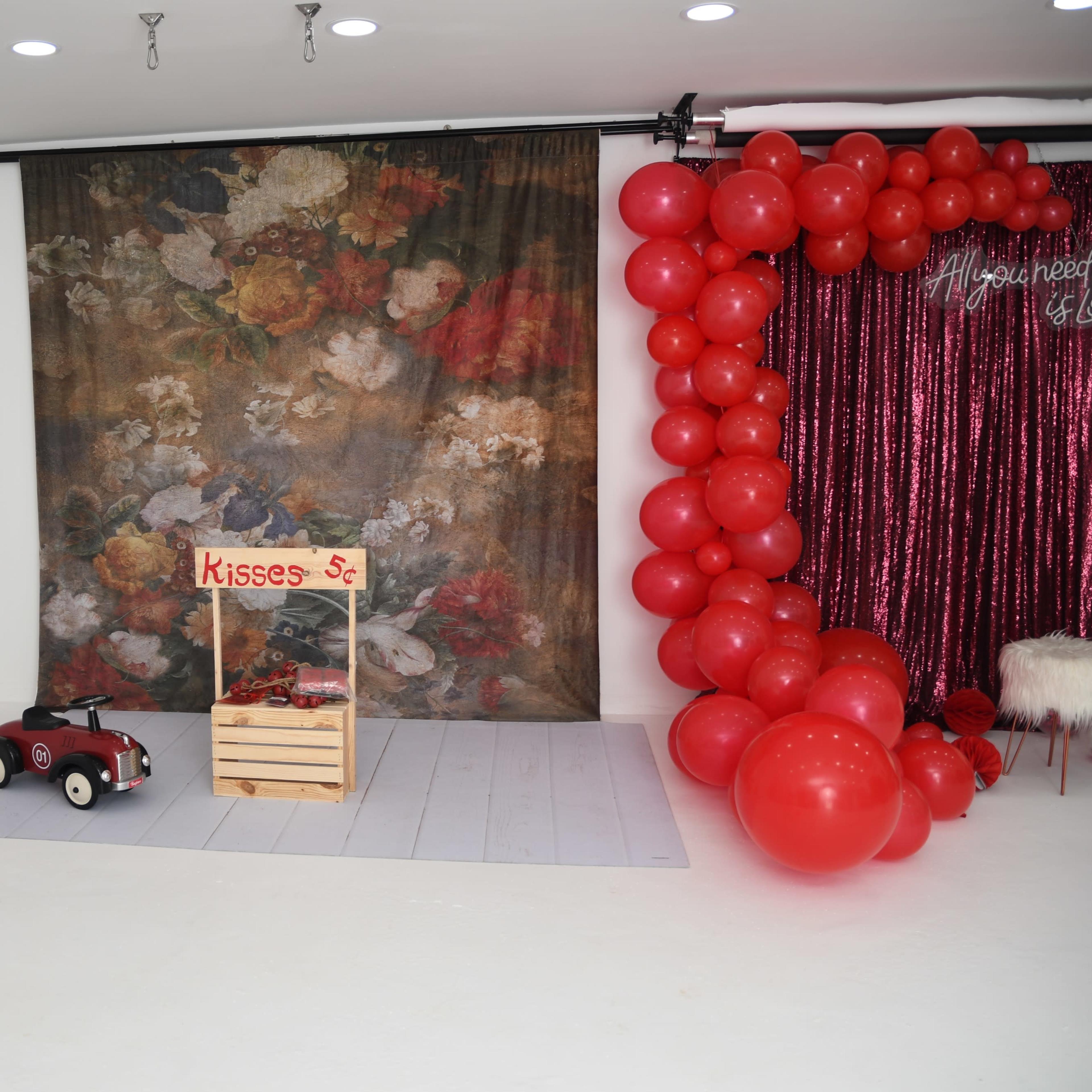 A well-decorated studio space with a white floor, a bench, greenery, balloons, a glittery backdrop, and a piano.