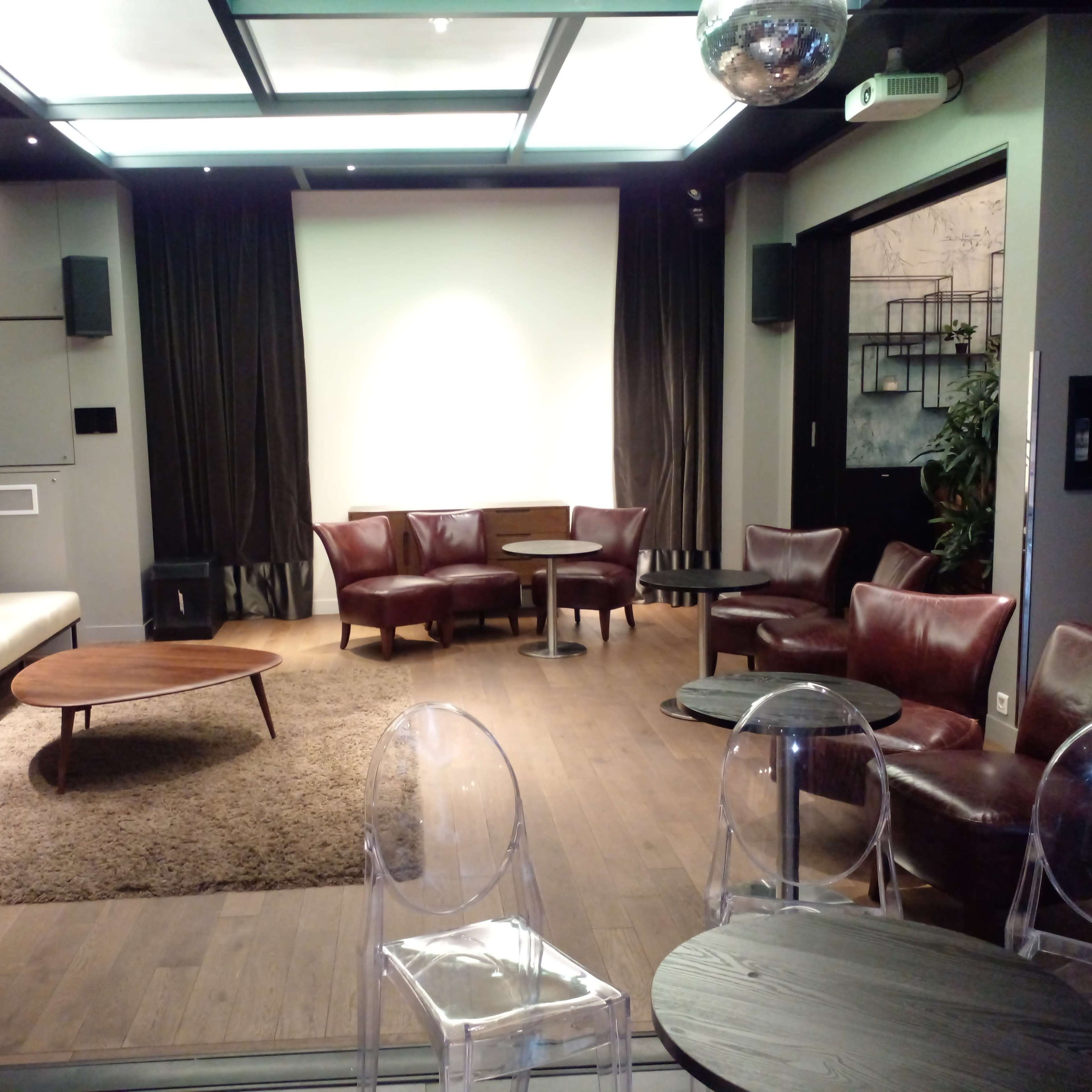 A modern lounge area with a white sofa, brown leather chairs, a wooden coffee table, and transparent chairs on a carpeted floor, illuminated by overhead lighting.