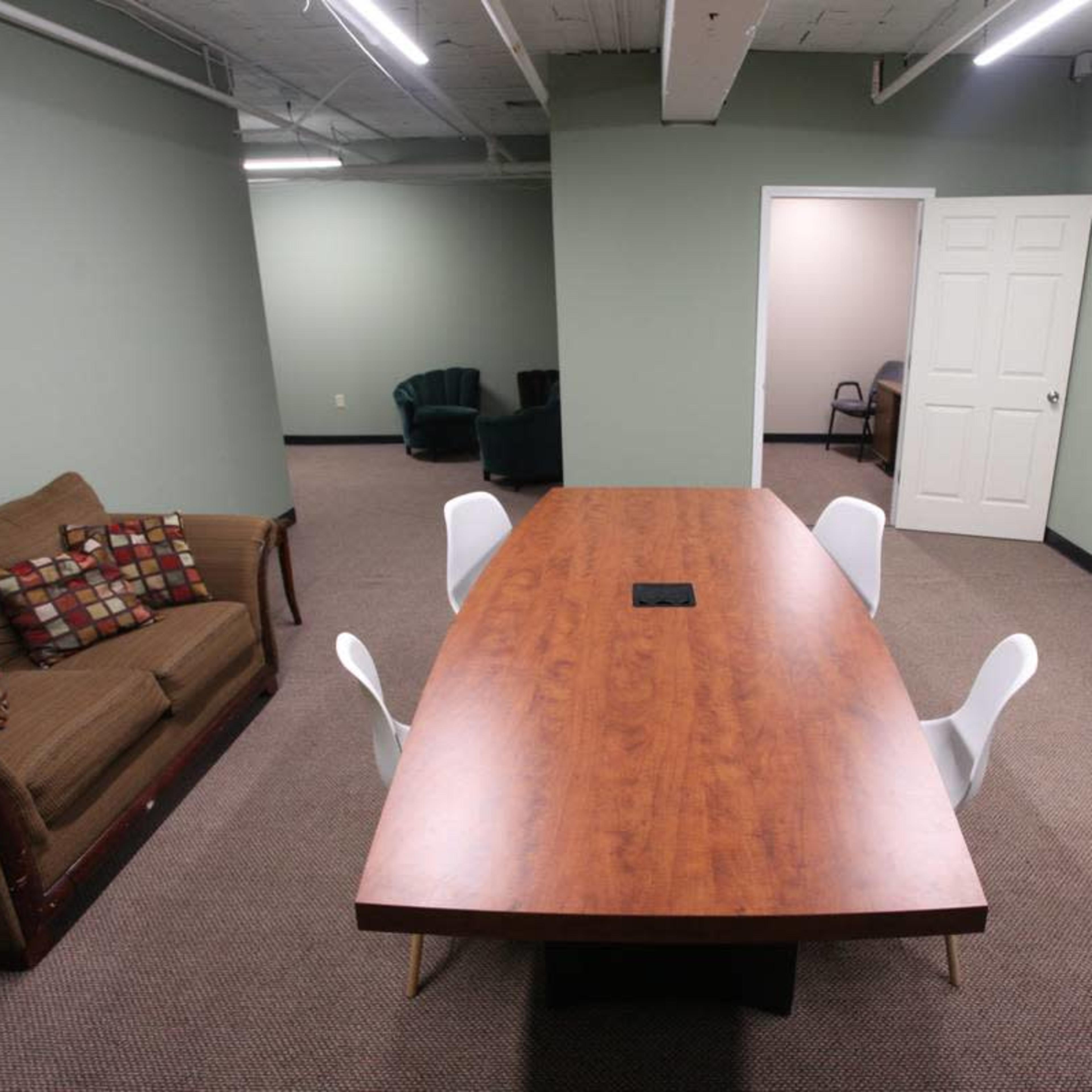 A conference room features a large wooden table surrounded by four white chairs, with a brown couch in the foreground and green walls.
