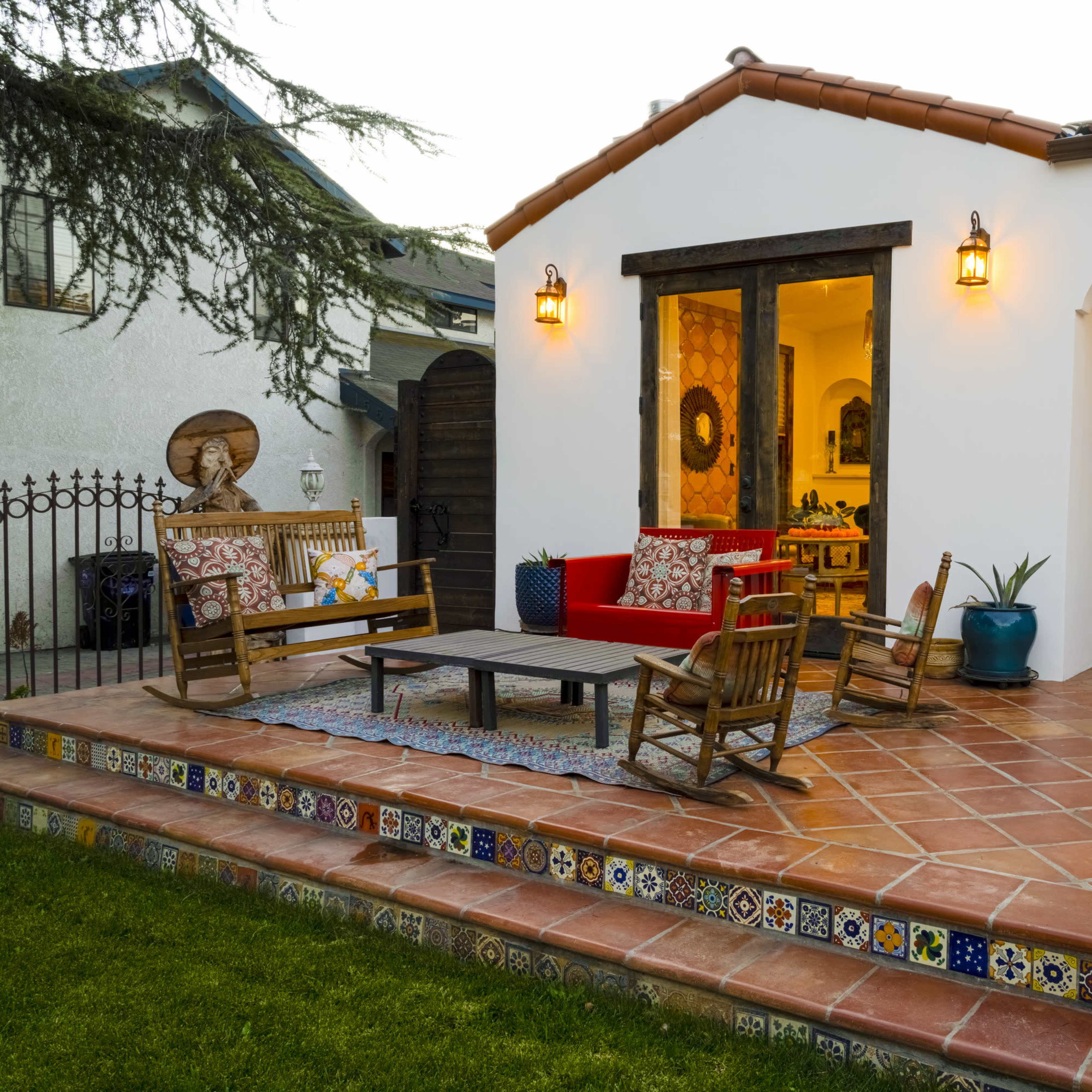 A cozy outdoor patio with terracotta tiles, wooden furniture, and decorative tiles along the steps, leading to a white stucco house with a wooden door and wall lights.