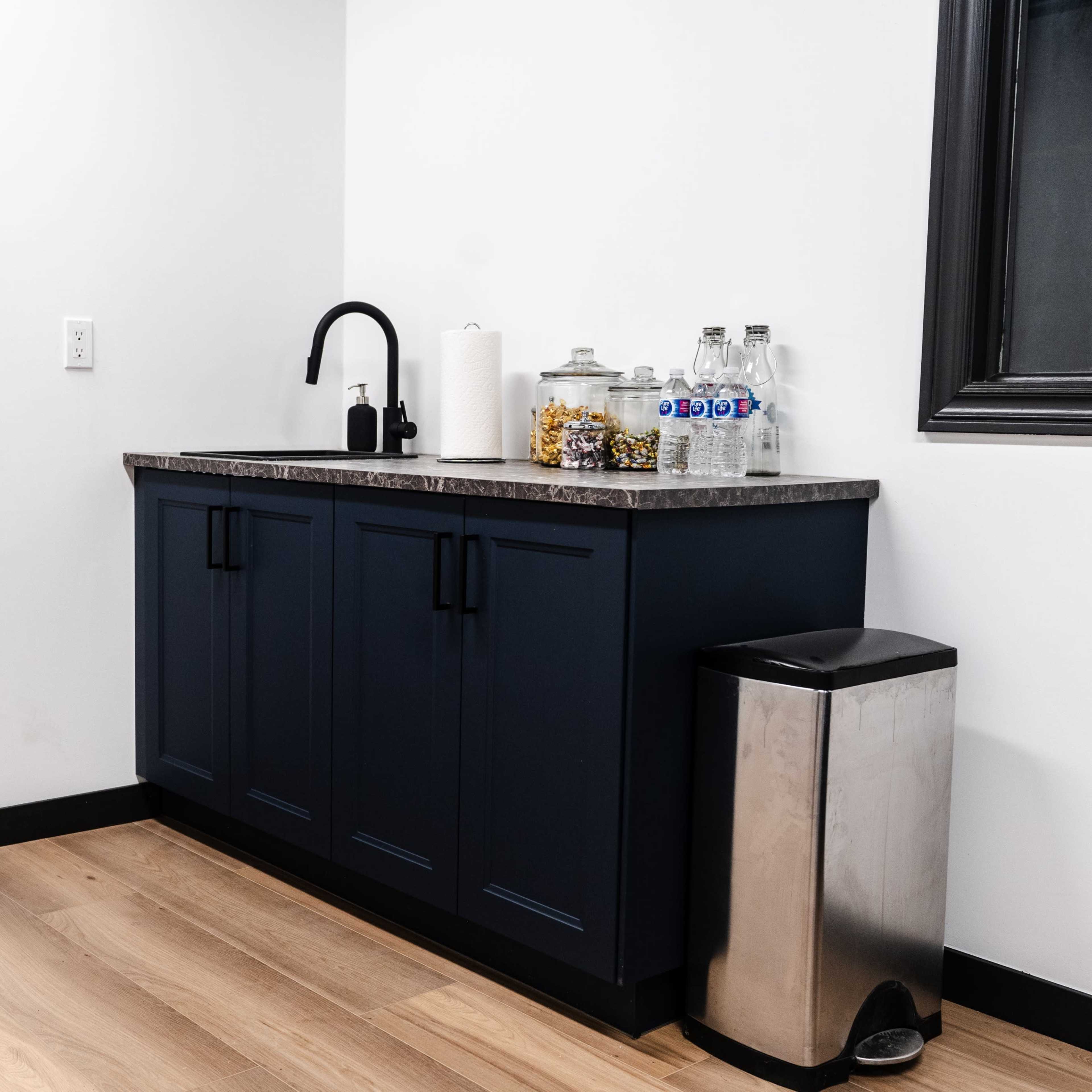 A kitchenette area featuring a dark cabinet with a countertop, a faucet, paper towels, translucent containers, and a trash can, along with wooden flooring and walls painted white.