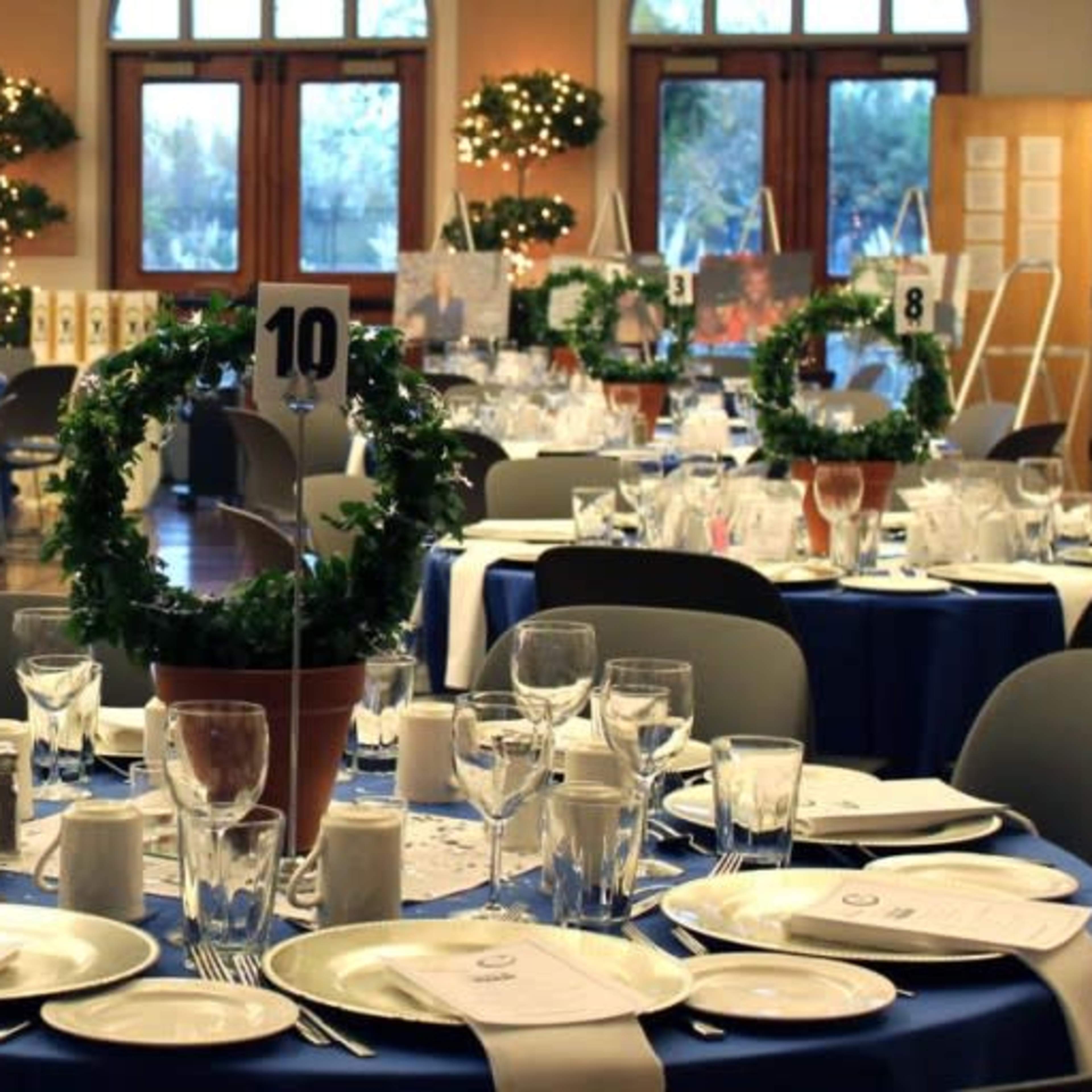 A banquet hall arranged with round tables set for dining, each featuring blue tablecloths, plates, glassware, and decorative centerpieces.