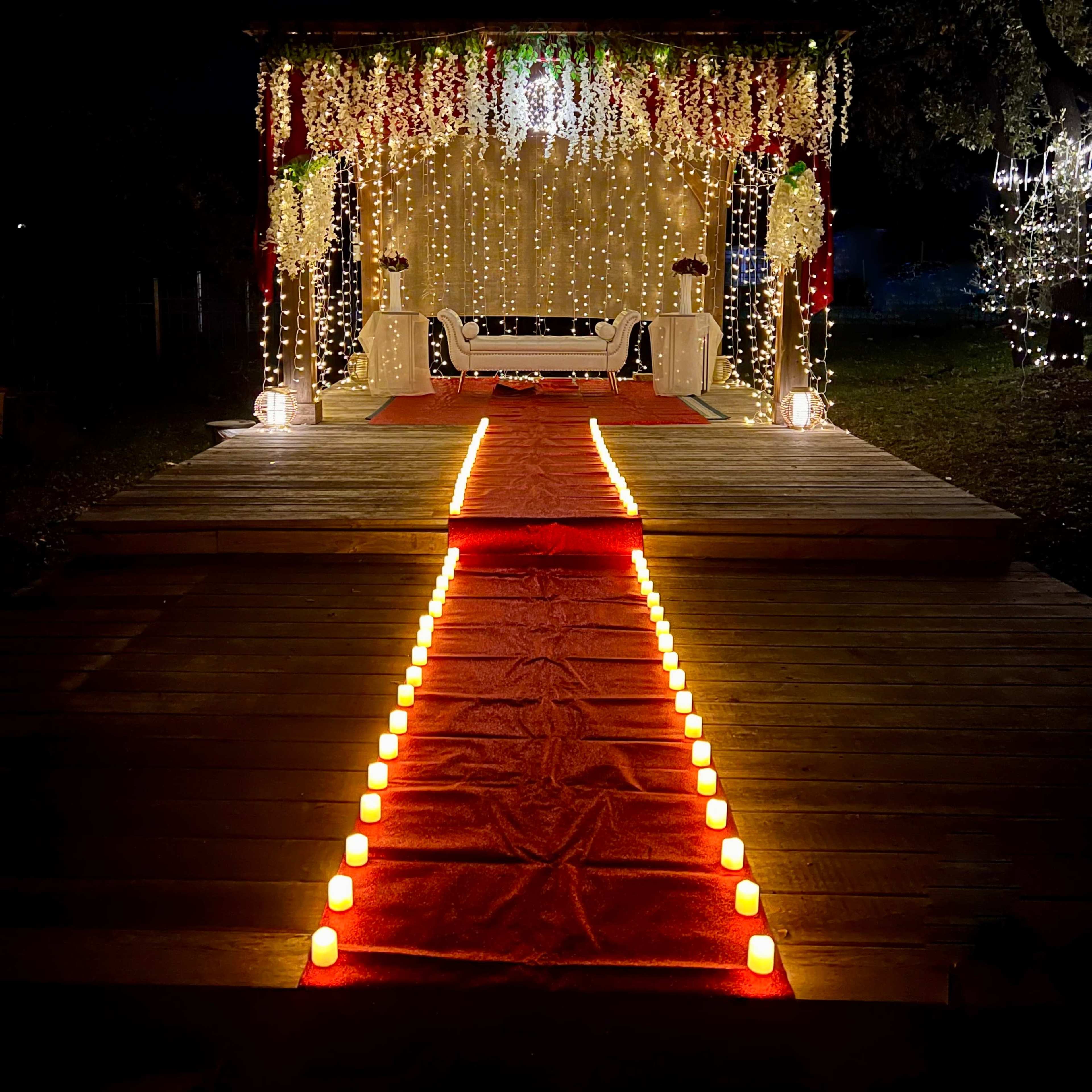 A decorated outdoor stage with a red carpet lined by candles and surrounded by string lights and floral arrangements.