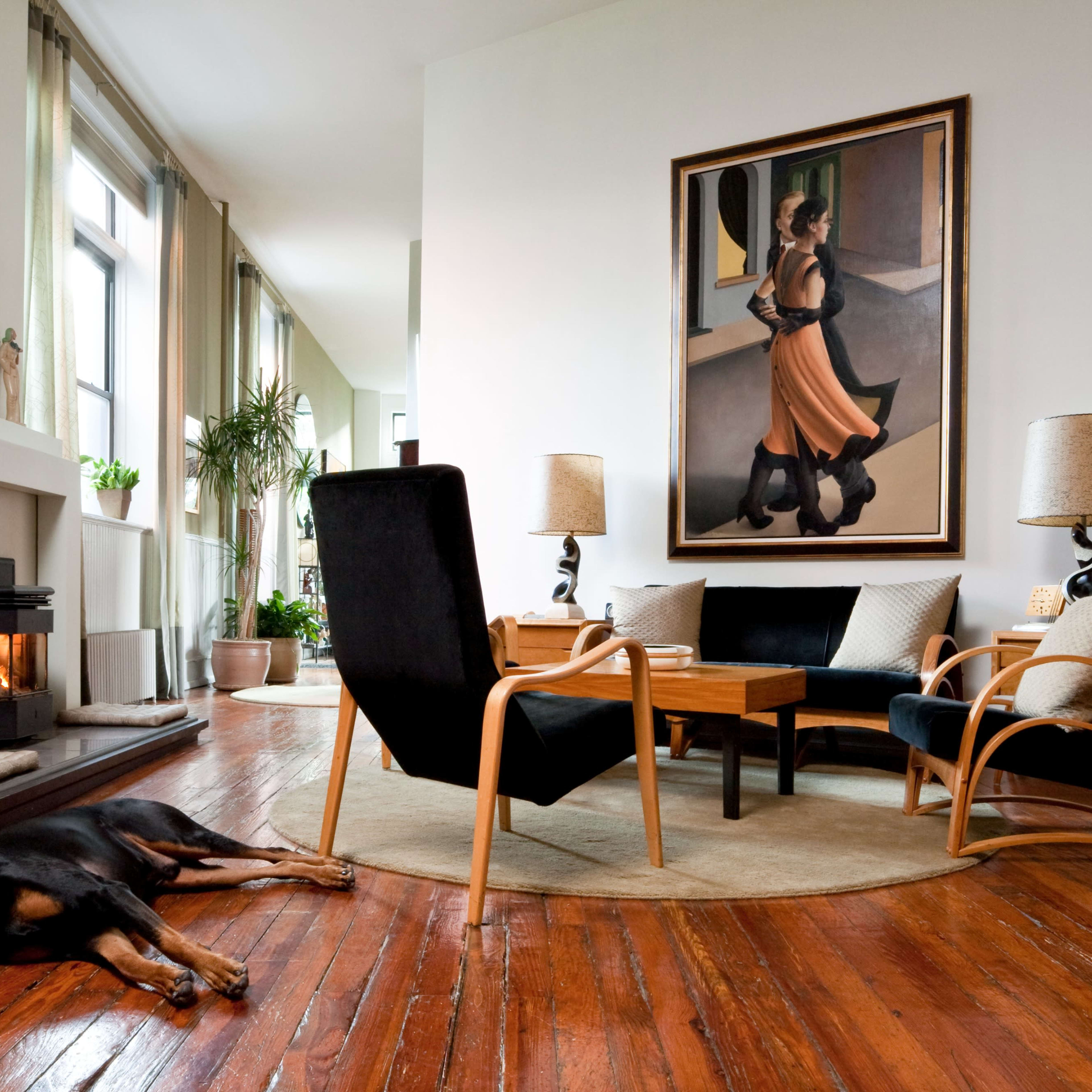 A spacious living room features a painting of a woman on the wall, a coffee table surrounded by two armchairs, and a dog resting on the wooden floor.