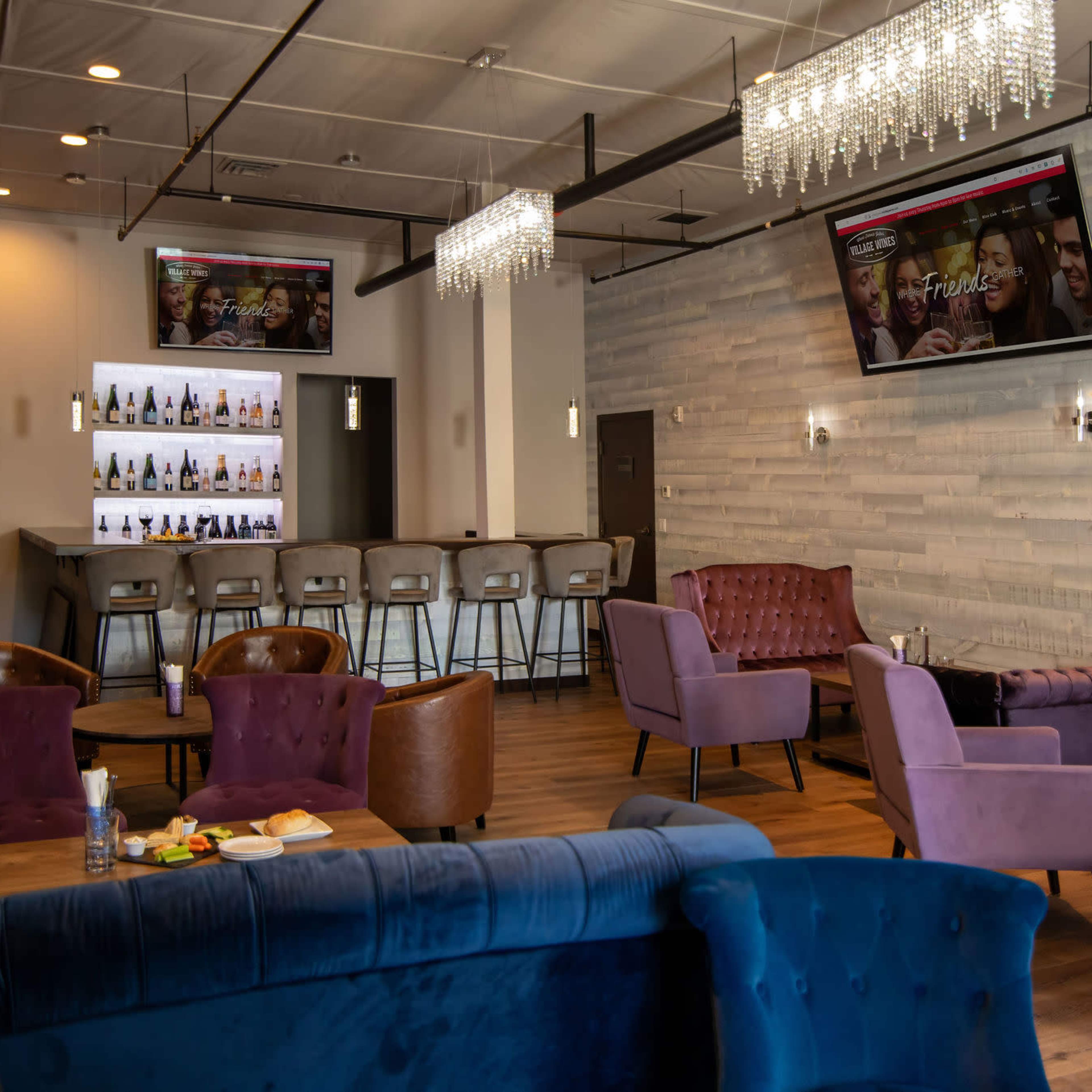A modern lounge area featuring plush seating, a bar with illuminated shelves, and two large screens displaying content.