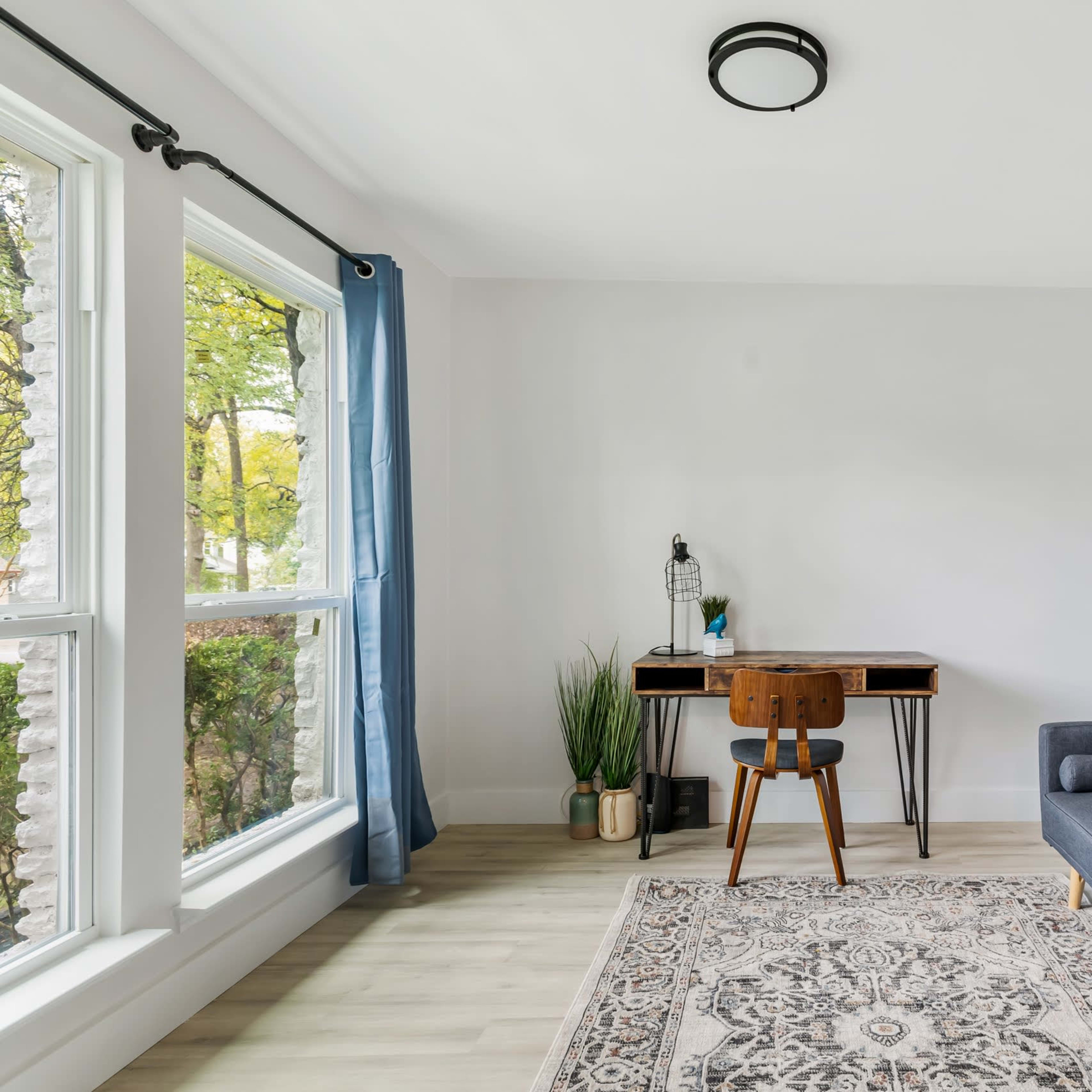 The room features a gray sofa, a wooden desk with a chair, a patterned rug, and large windows with light blue curtains.