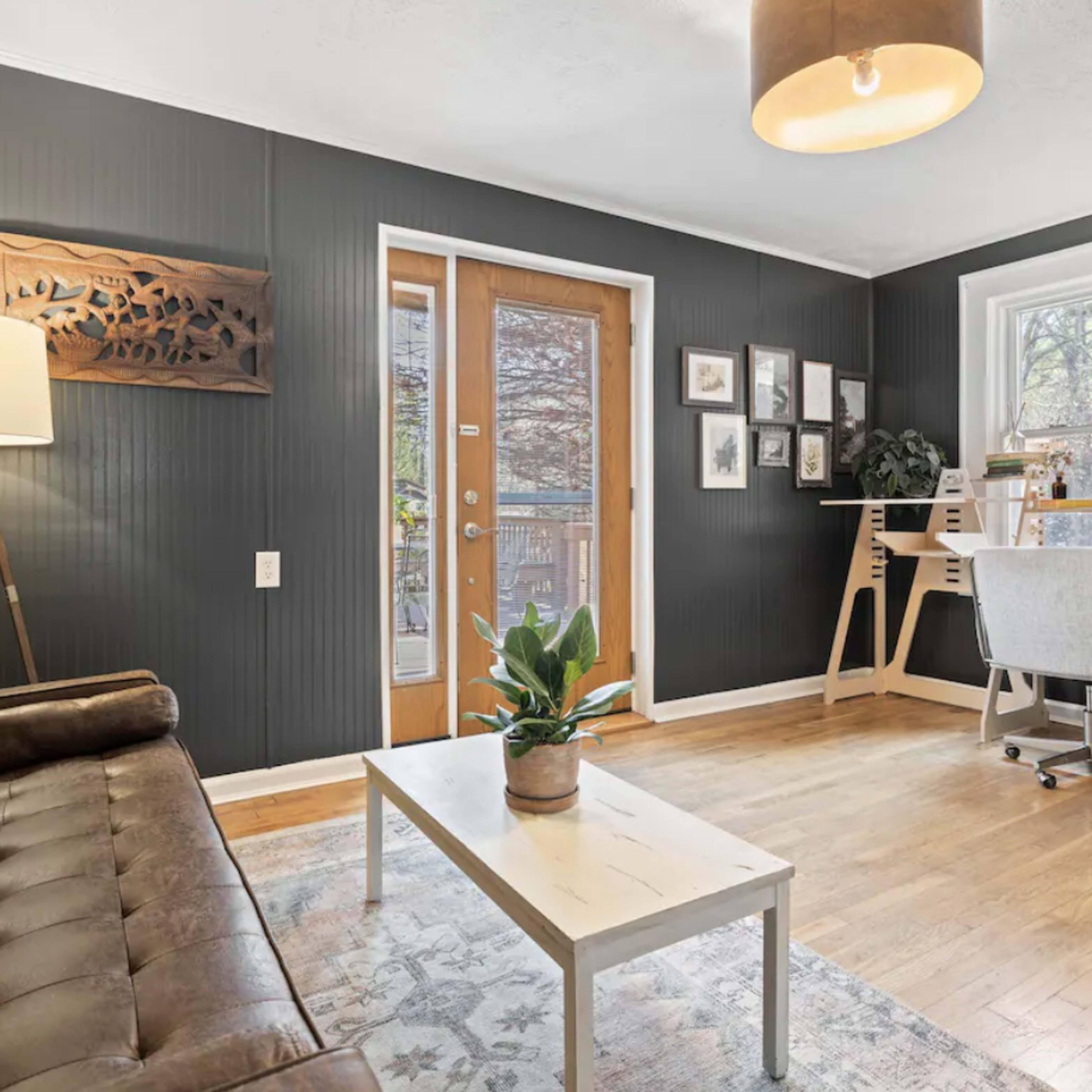 A living room with dark wood-paneled walls, featuring a brown leather sofa, a white coffee table, and a workspace with a desk and chair near a door leading outside.