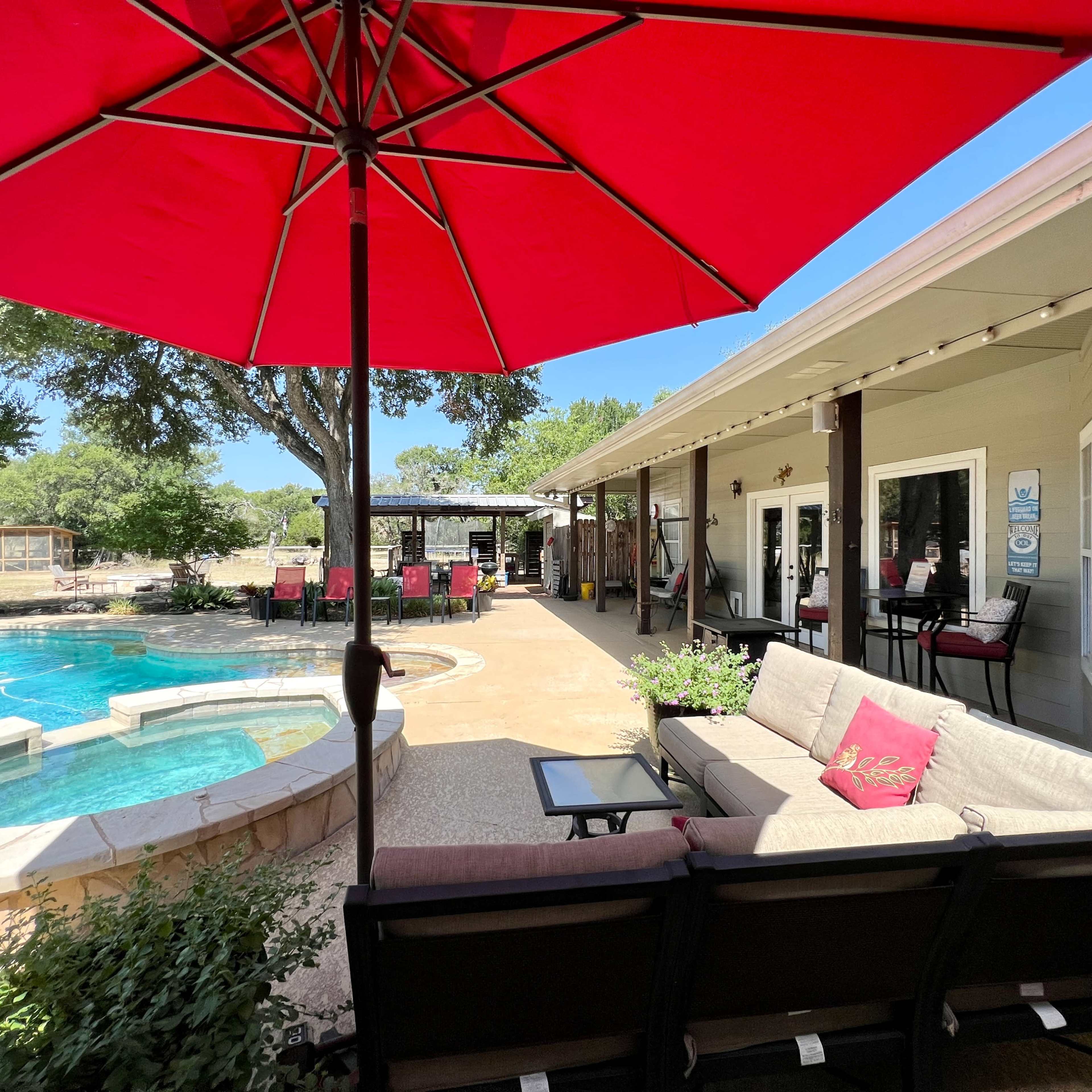 A cozy outdoor seating area with a red umbrella overlooks a swimming pool and a nearby patio with lounge chairs.