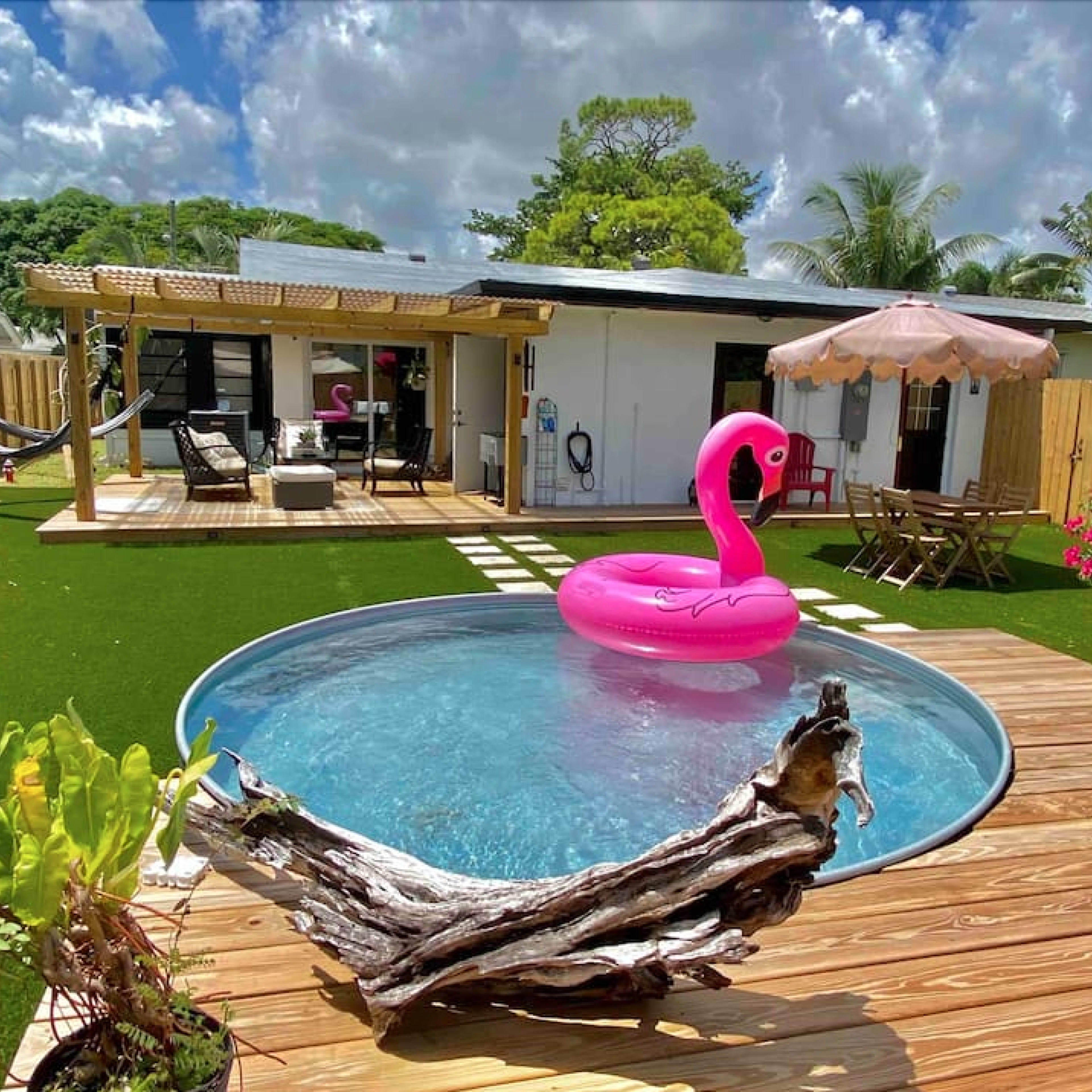 A backyard features a round swimming pool with a pink flamingo float, surrounded by wooden decking, lounge furniture, and a shaded seating area.