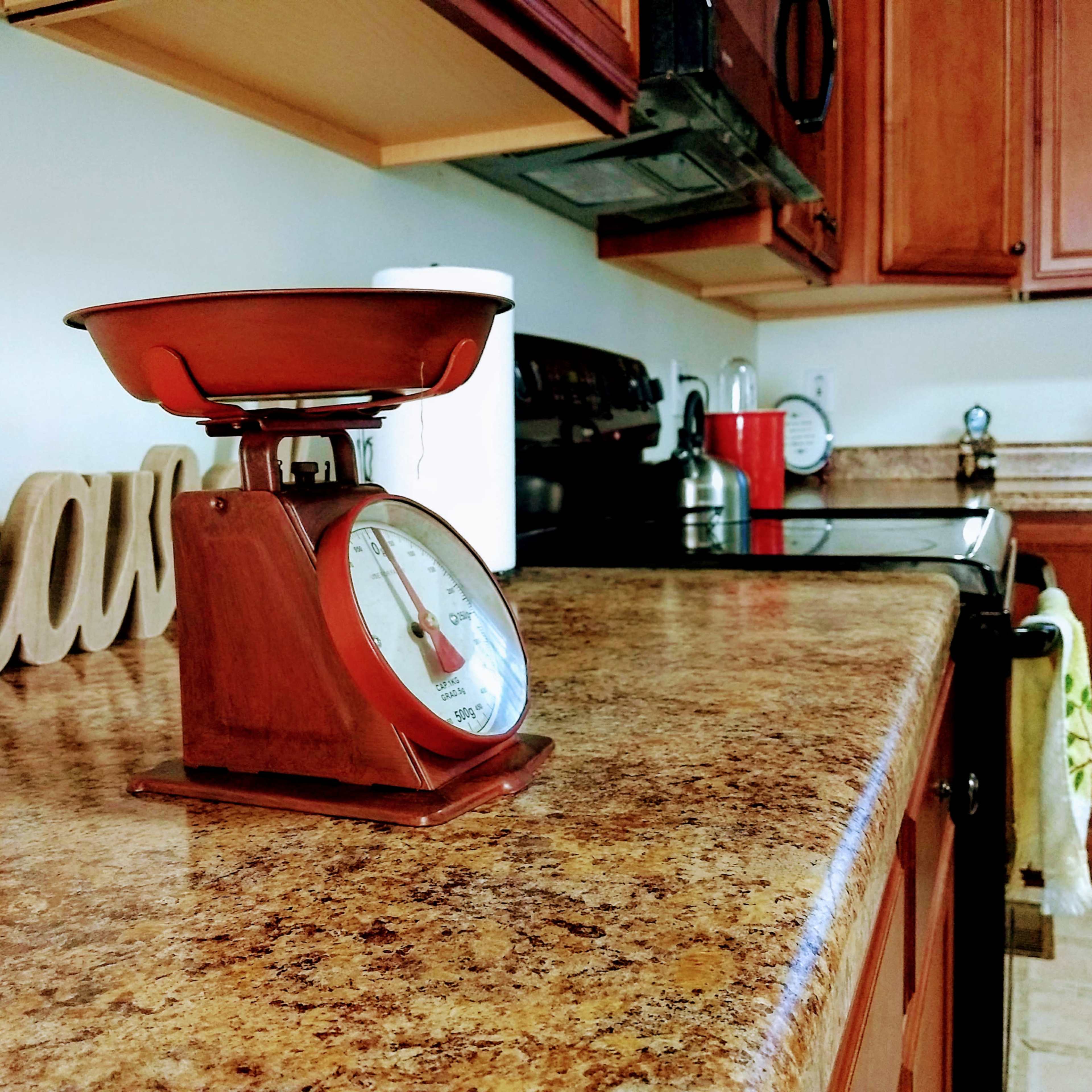 A vintage kitchen scale sits on a granite countertop in a well-equipped kitchen with wooden cabinets.