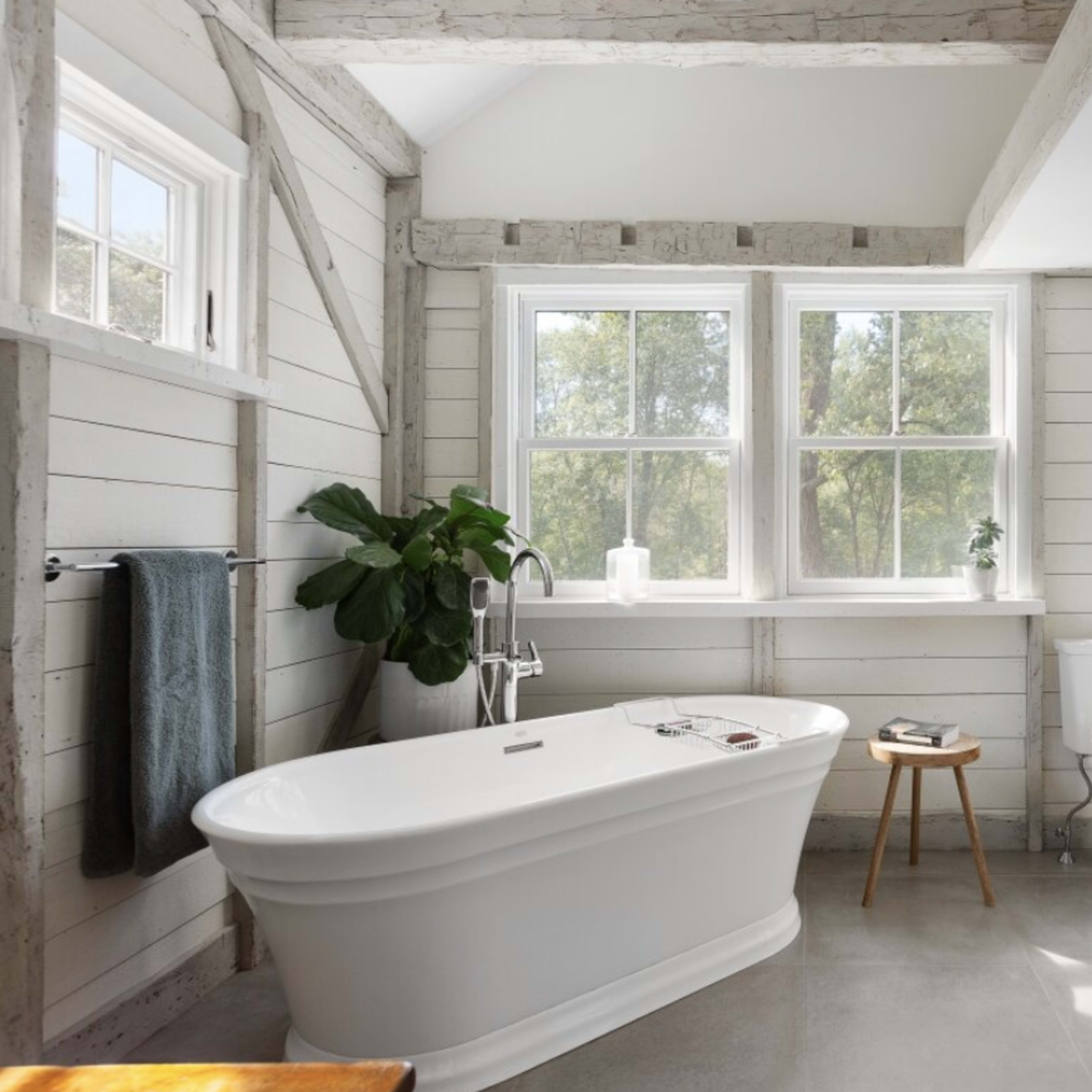 A bright bathroom with a freestanding white bathtub, a toilet, and large windows, featuring wooden accents and a potted plant.