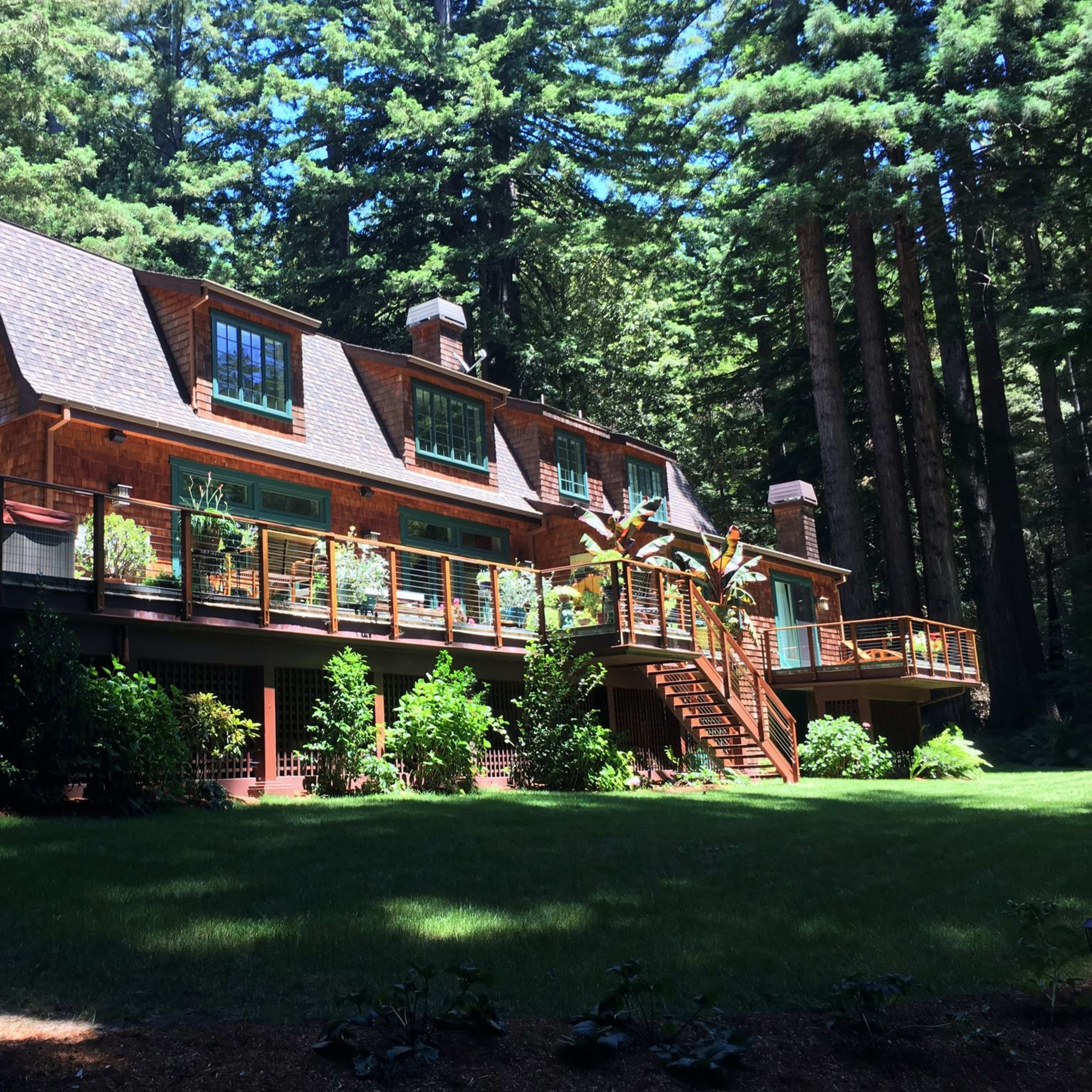 A large wooden house with multiple windows and a deck surrounded by tall green trees.
