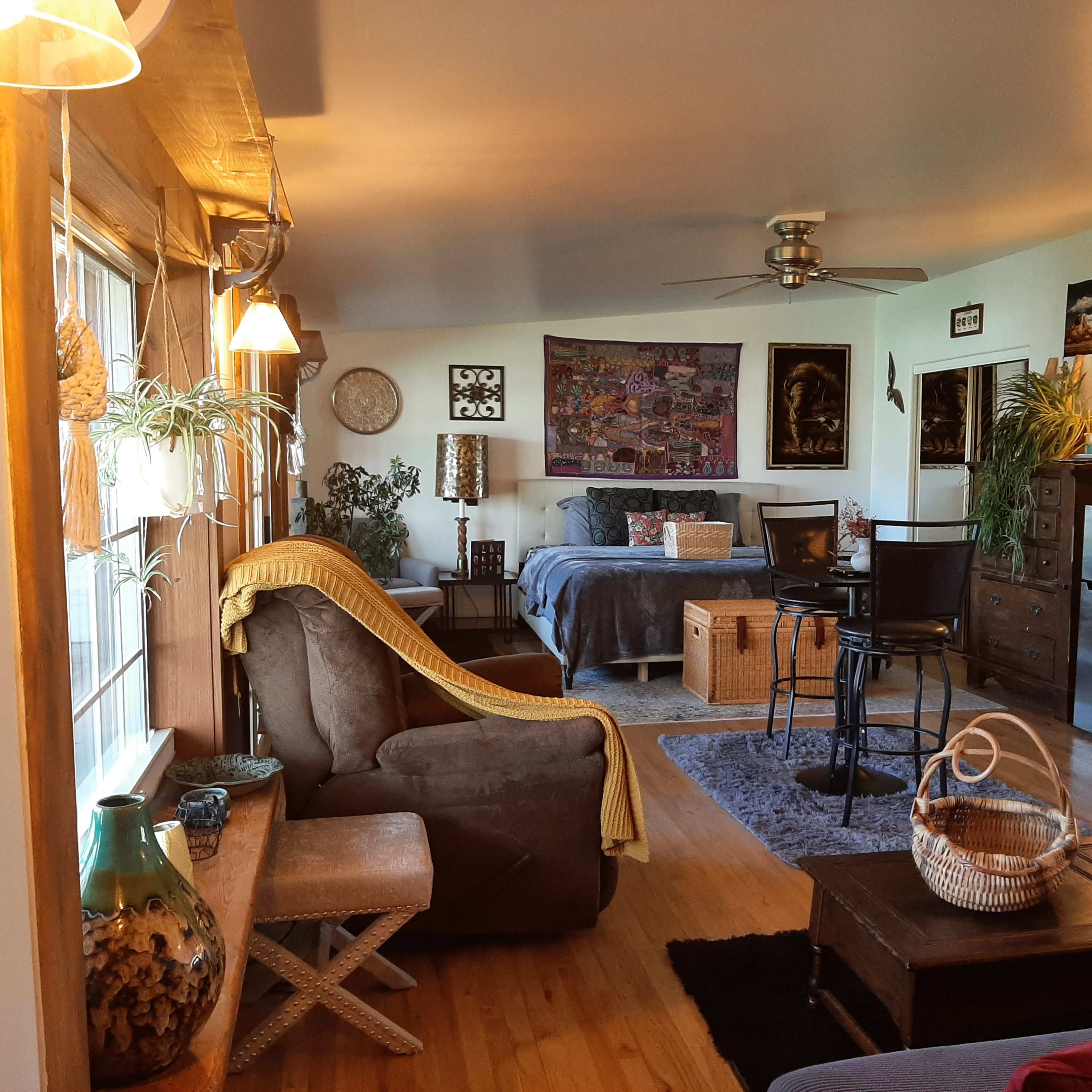 A spacious living area features a mix of seating, decorative plants, and artwork, with a dining area and kitchen visible in the background.