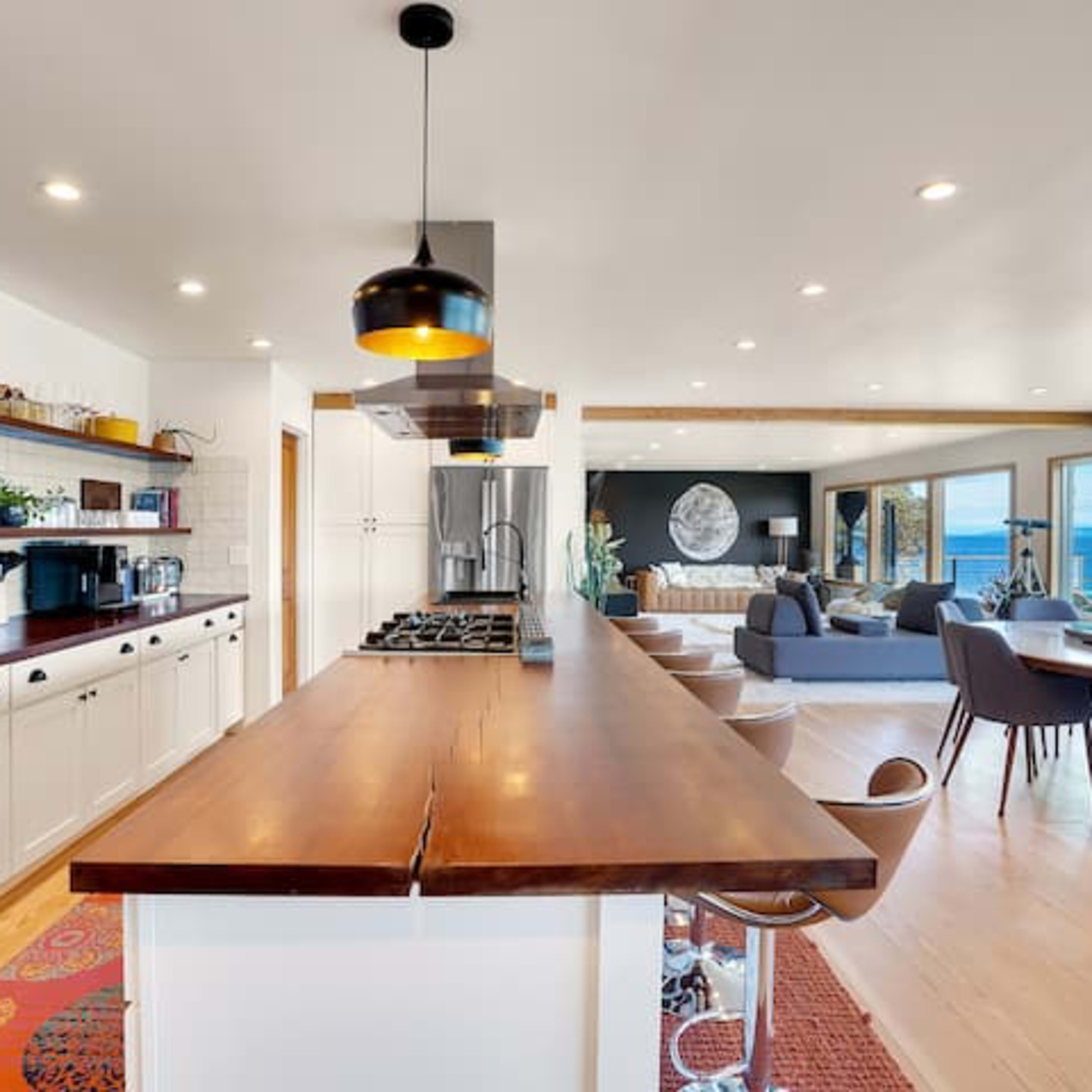 A modern kitchen with a central wooden island, featuring a stove, surrounded by a light-filled open living area that includes a dining area and seating.
