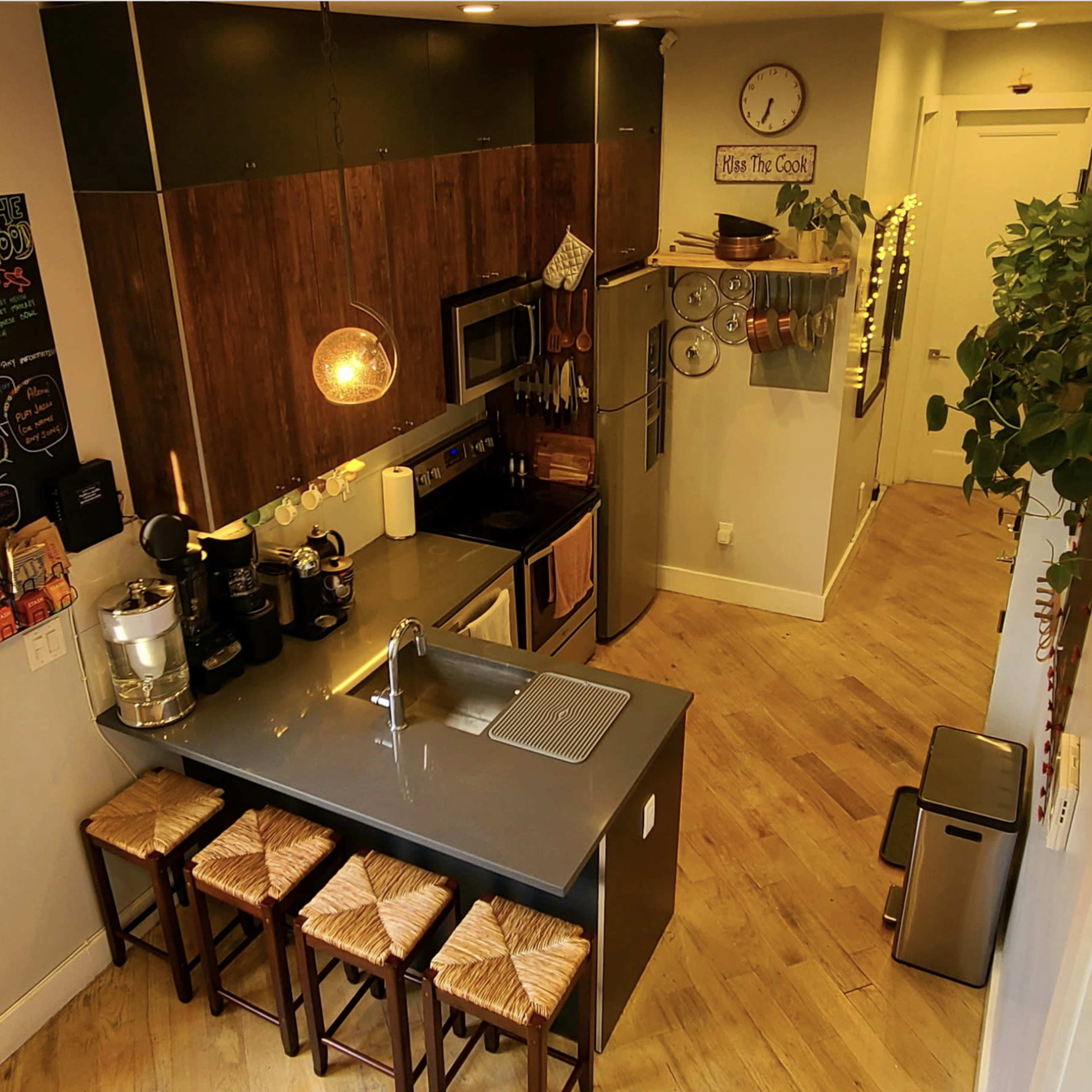 A modern kitchen with an island, equipped with stainless steel appliances, wooden cabinetry, and three stools, surrounded by plants.