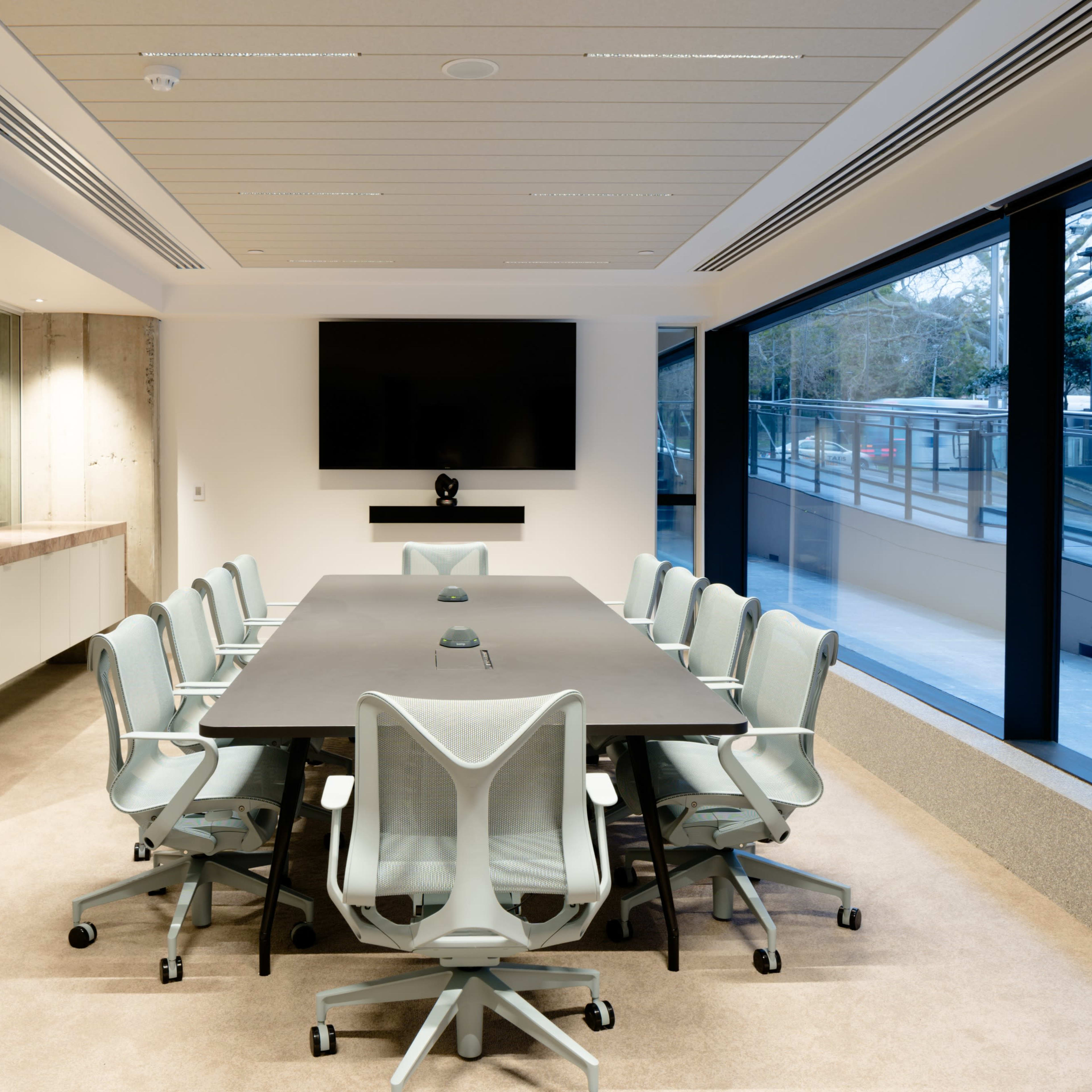 A modern conference room features a large rectangular table surrounded by ergonomic chairs, with a large screen mounted on the wall.