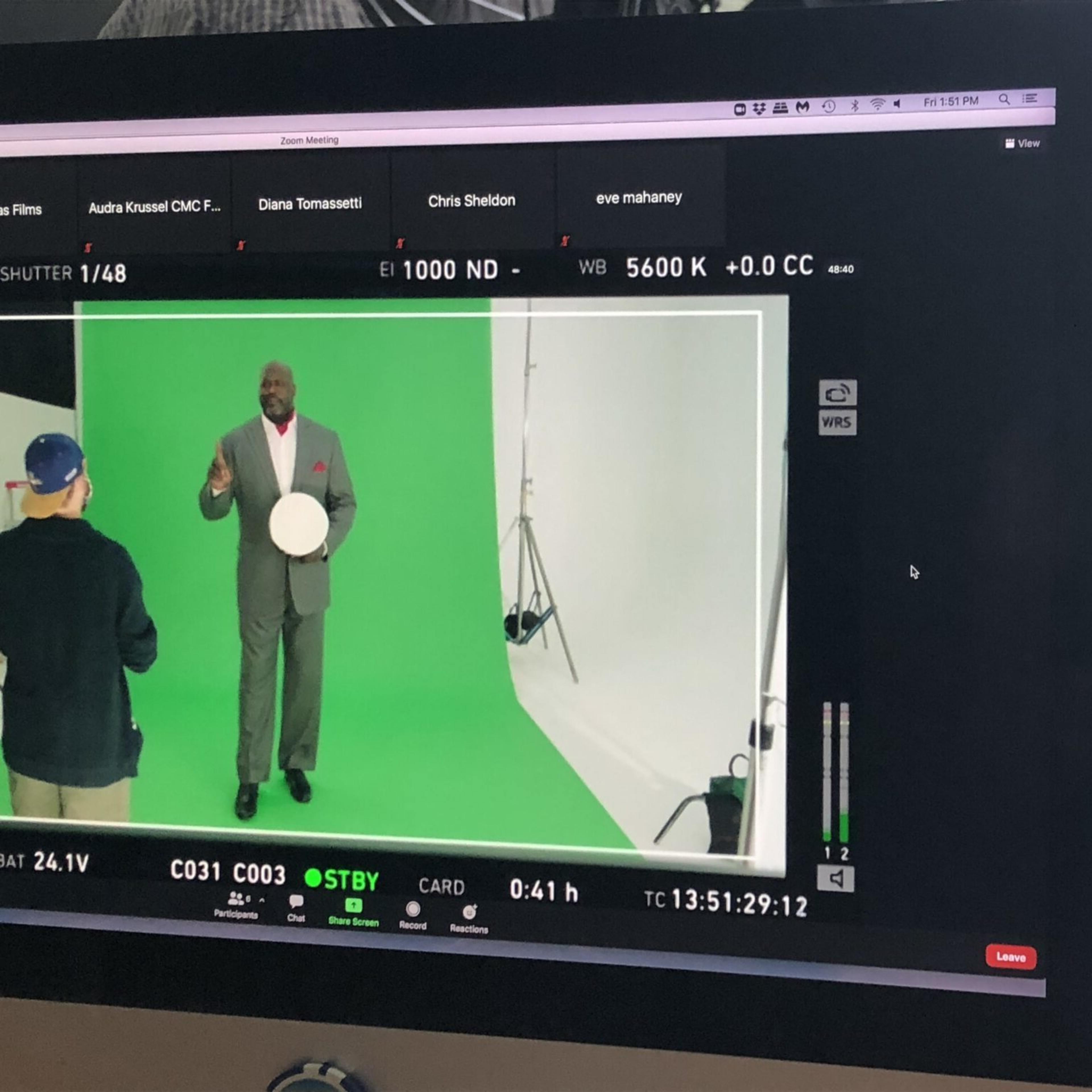 A man in a gray suit stands in front of a green screen while holding a circular object, as he addresses a person off-camera.