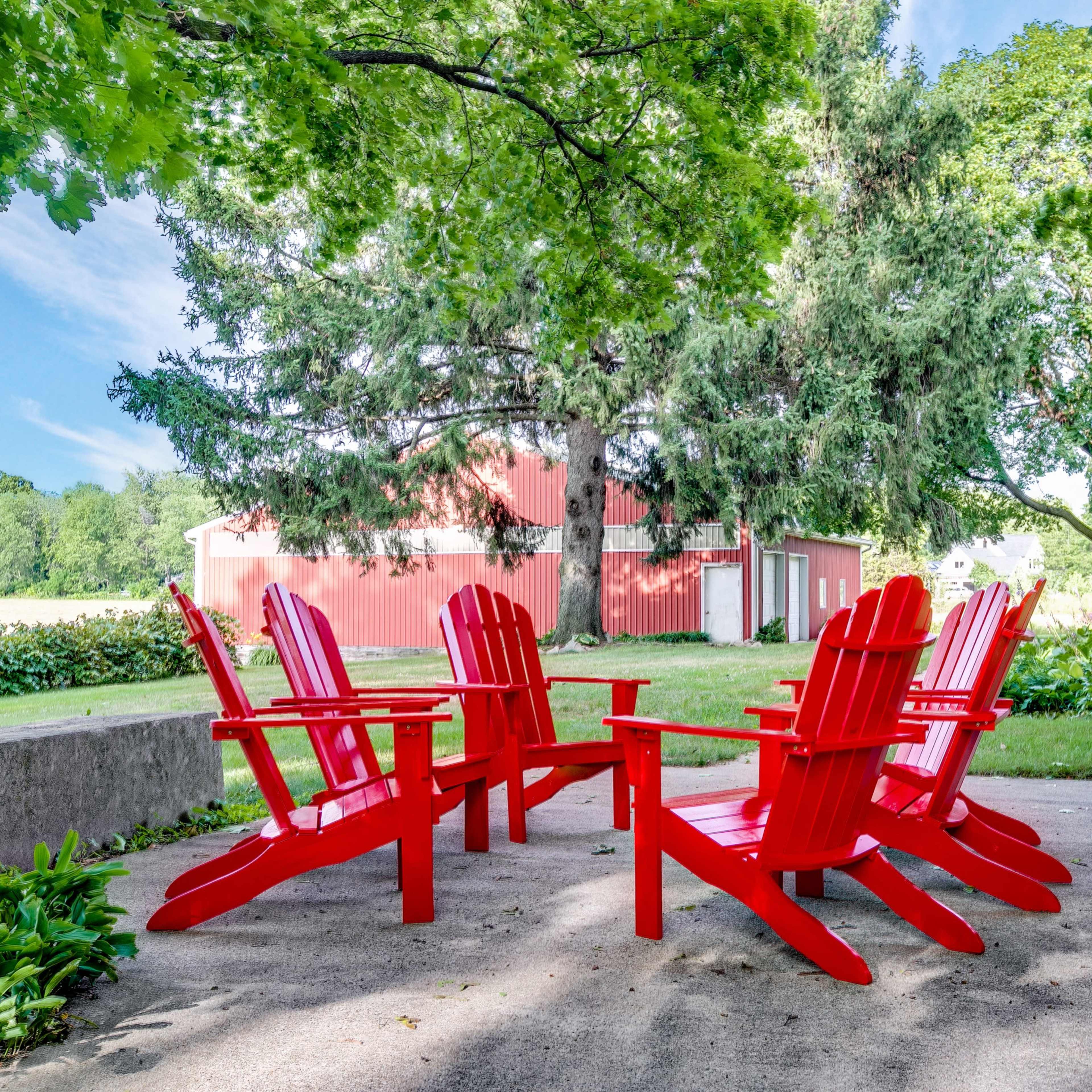 A circle of six red adirondack chairs is arranged on a concrete patio under large trees near a red barn and house.