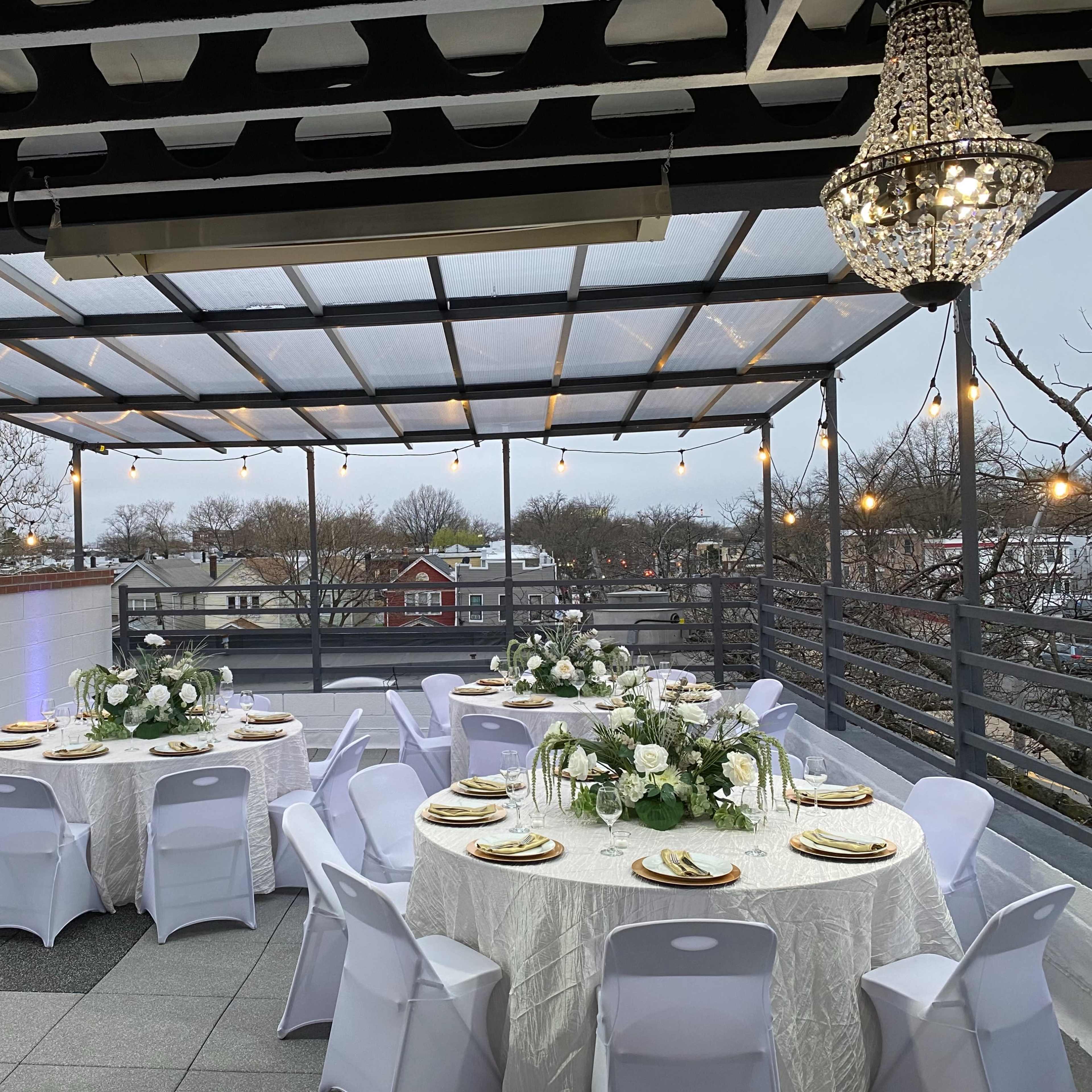 A rooftop event space with tables set for dining, adorned with white tablecloths, floral centerpieces, and elegant chandeliers overhead, framed by string lights and a view of nearby buildings.