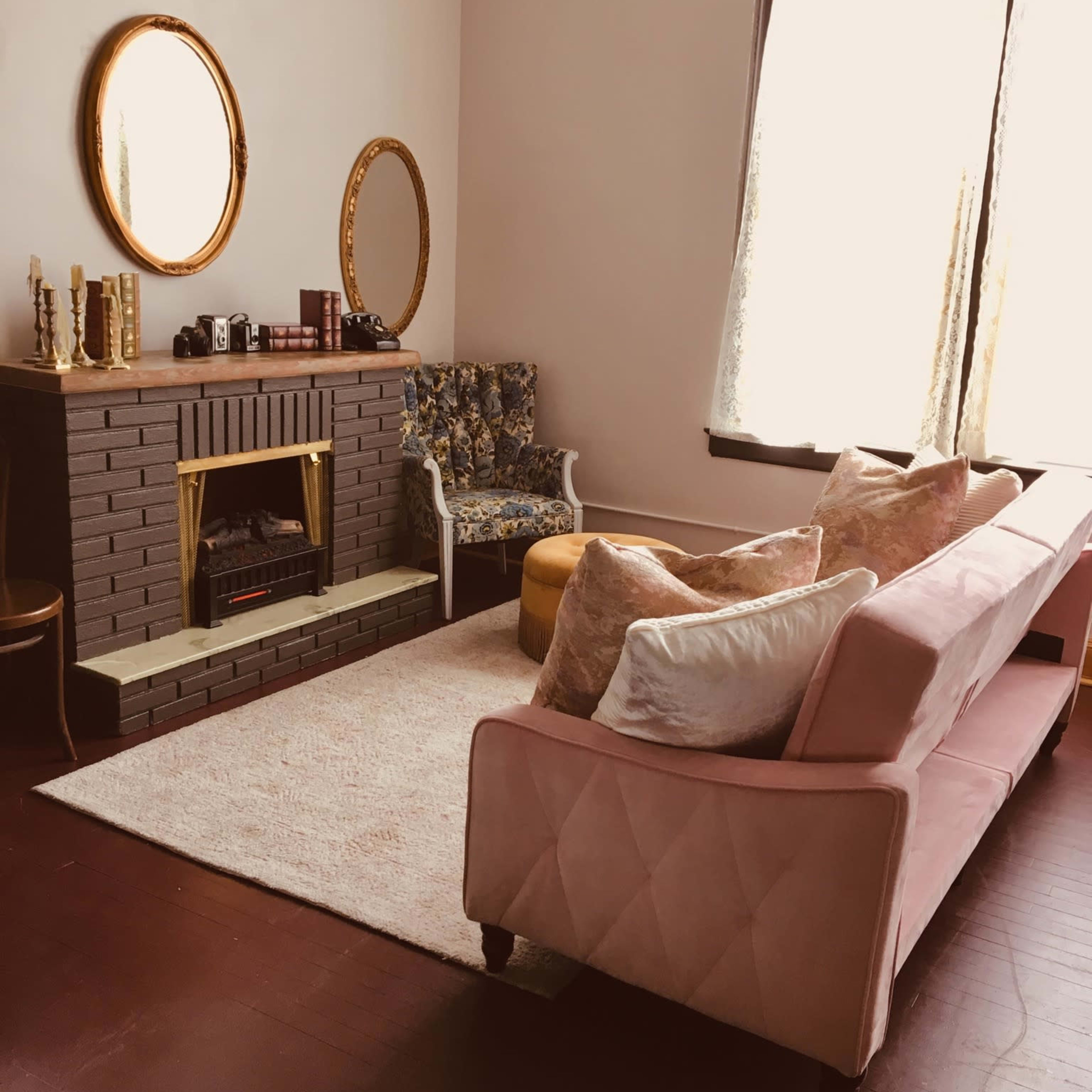A cozy living room with a pink couch, a patterned armchair, a gold-framed mirror, and a fireplace surrounded by decorative items.