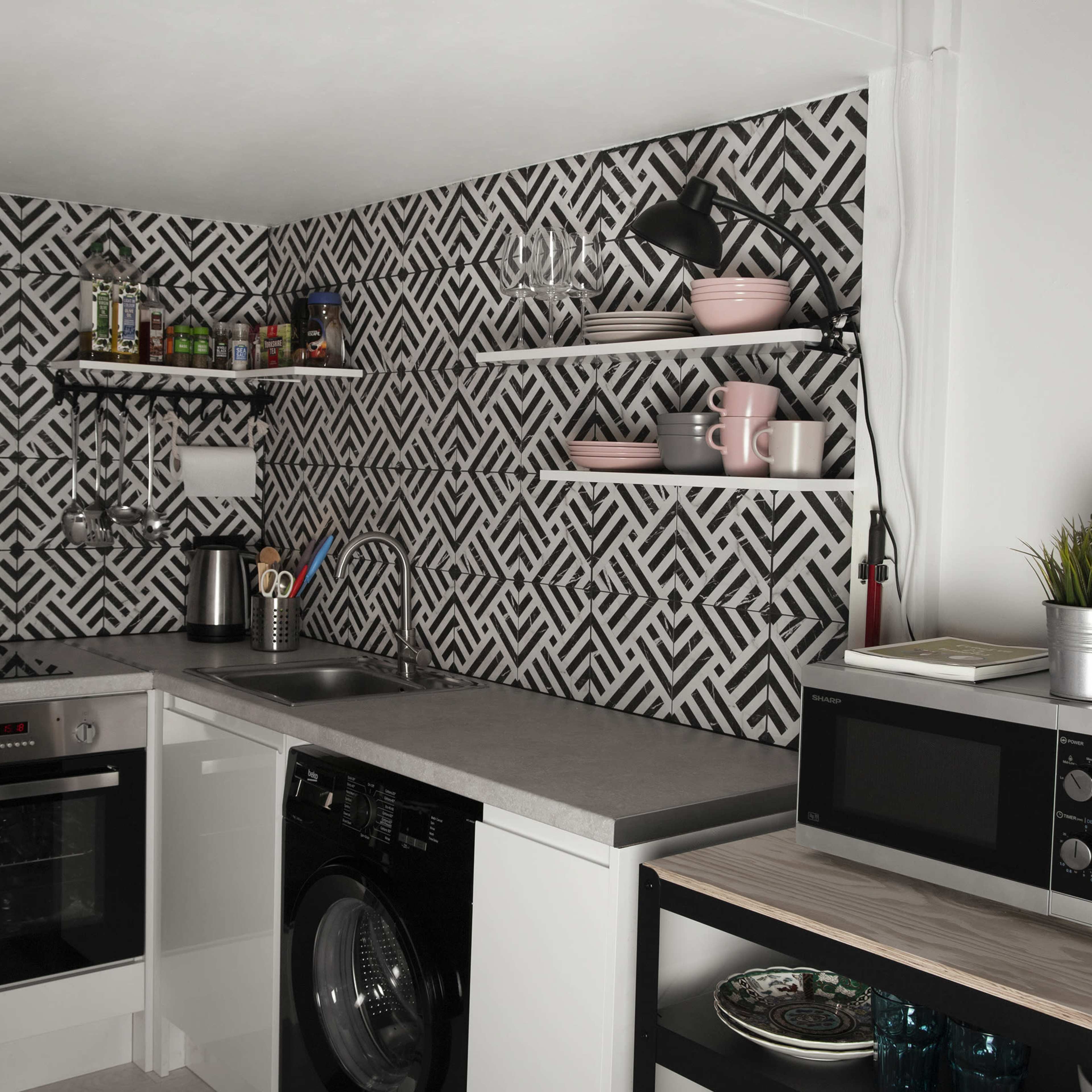 A modern kitchen features a geometric patterned wall, stainless steel appliances, and open shelving displaying pink dishware.