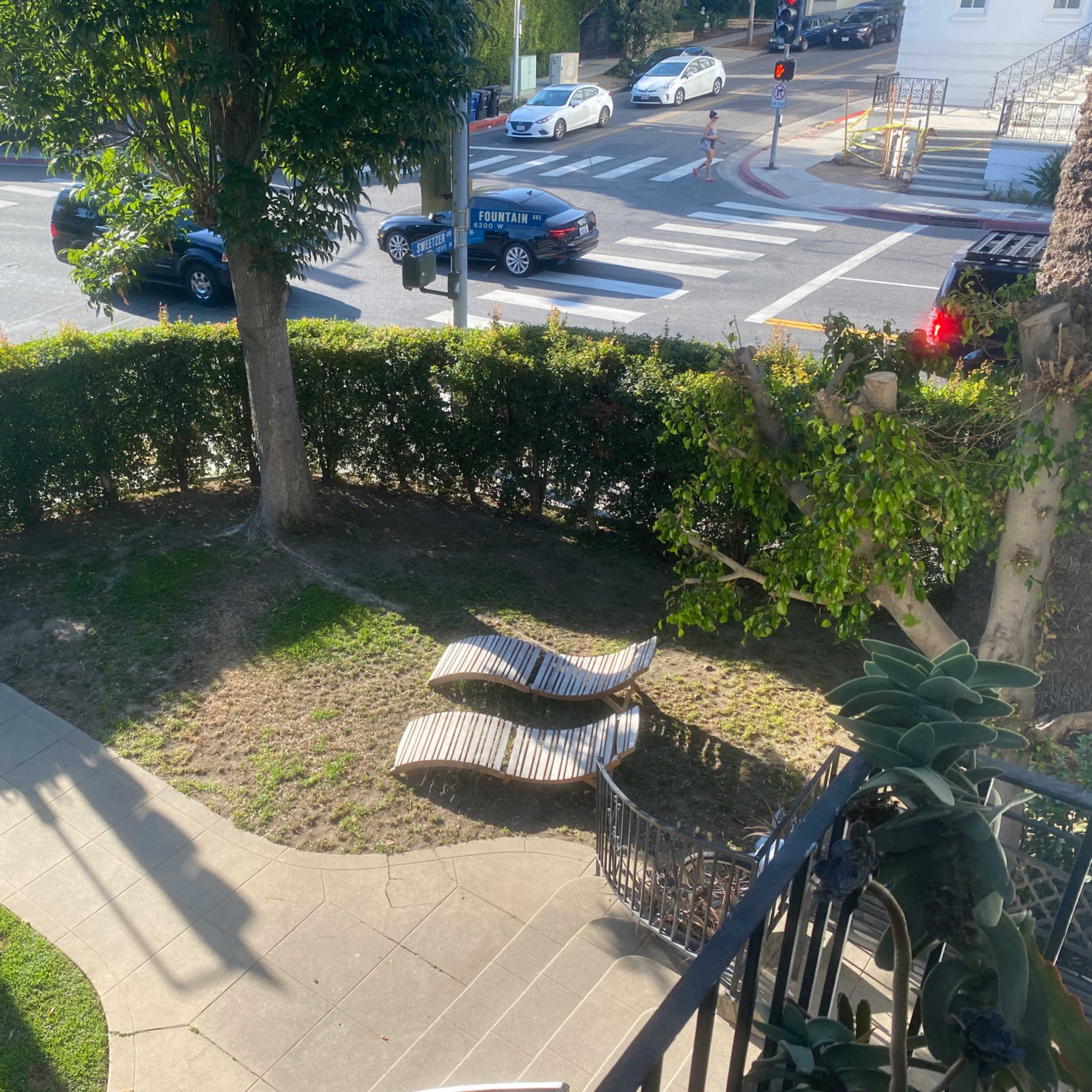 A small green area with two lounge chairs surrounded by a hedge, adjacent to a busy street with cars and traffic signals.