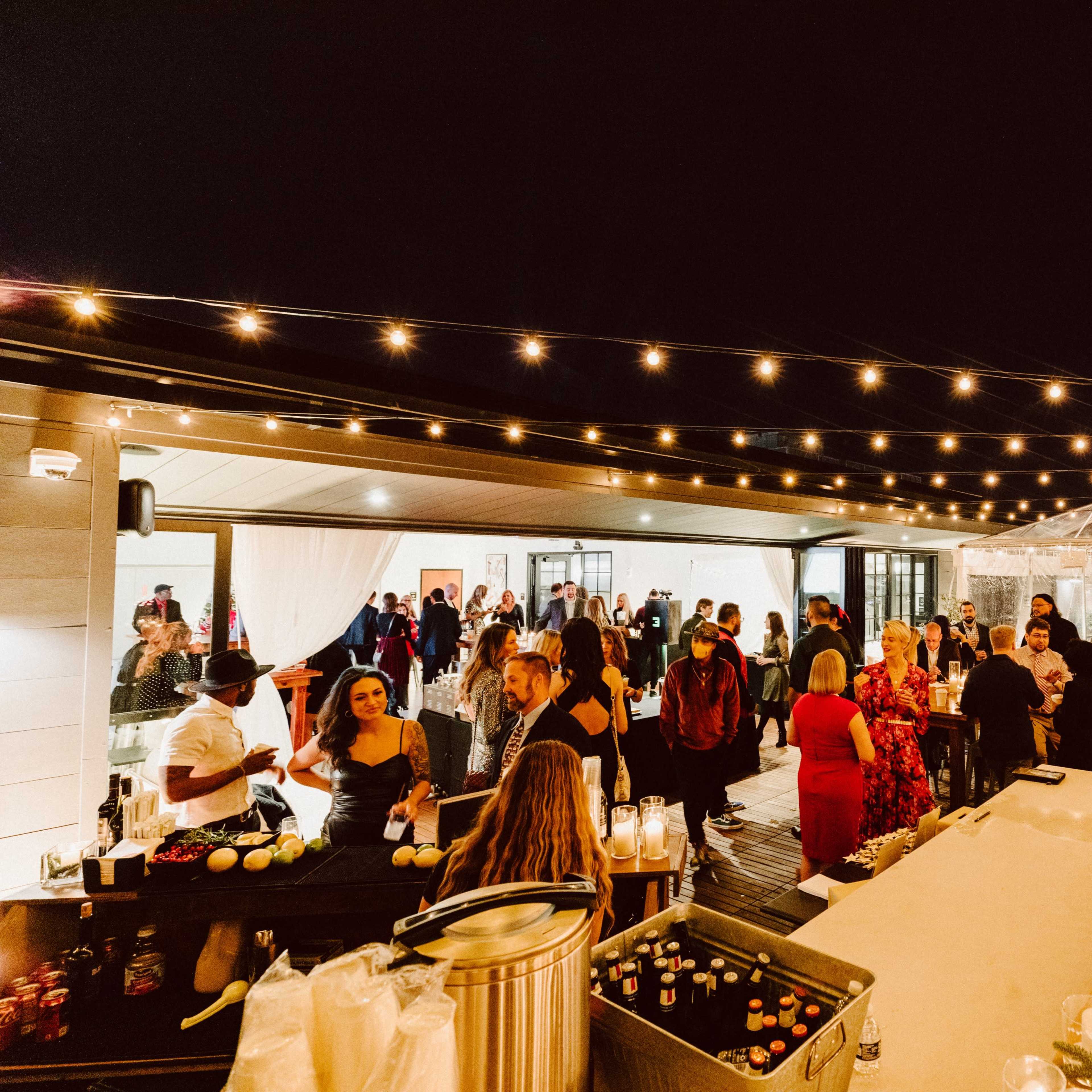 A large gathering takes place outdoors at night, featuring a bar area and string lights overhead, with guests mingling under a tent.