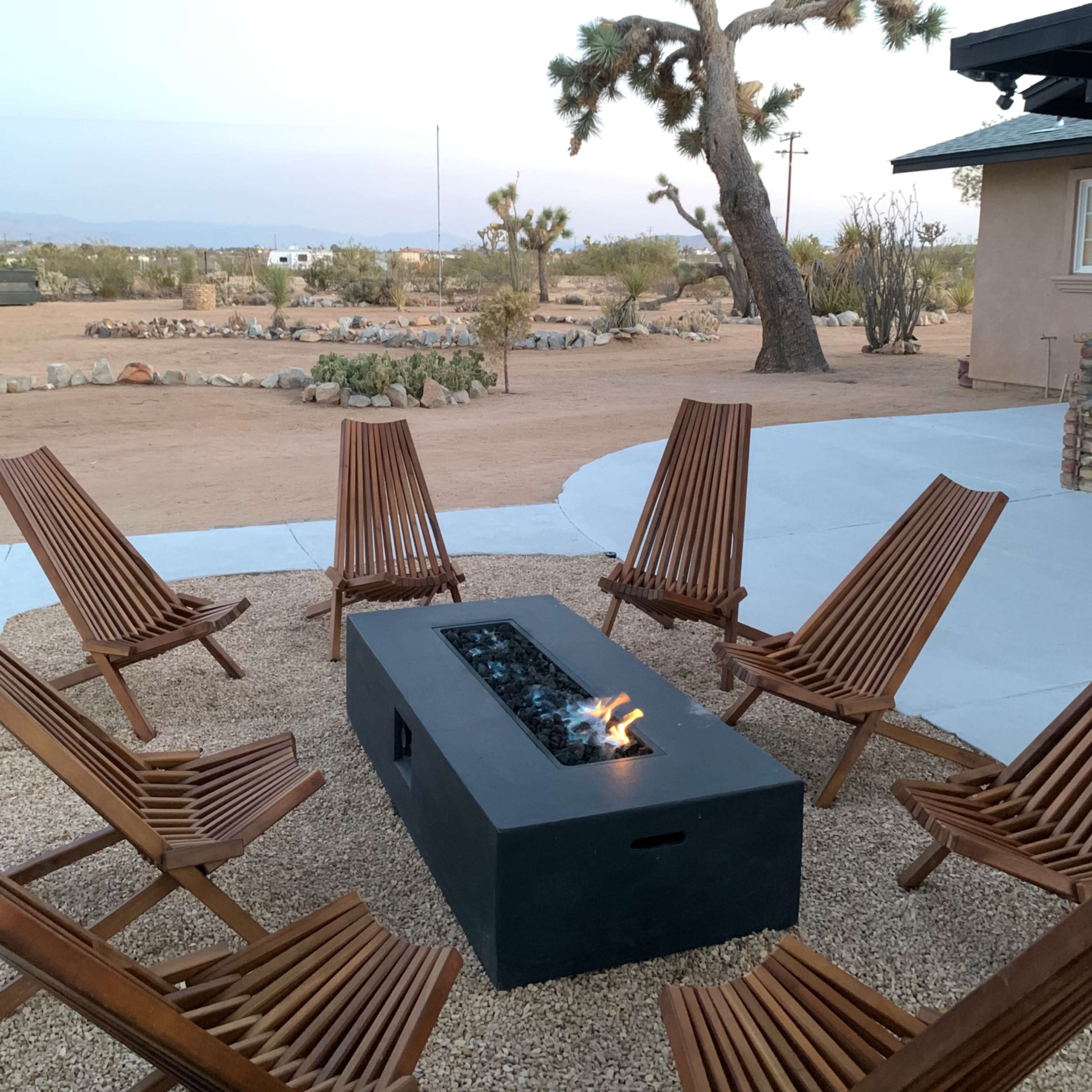 A fire pit surrounded by wooden chairs is situated on a gravel patio in a desert landscape with Joshua trees in the background.