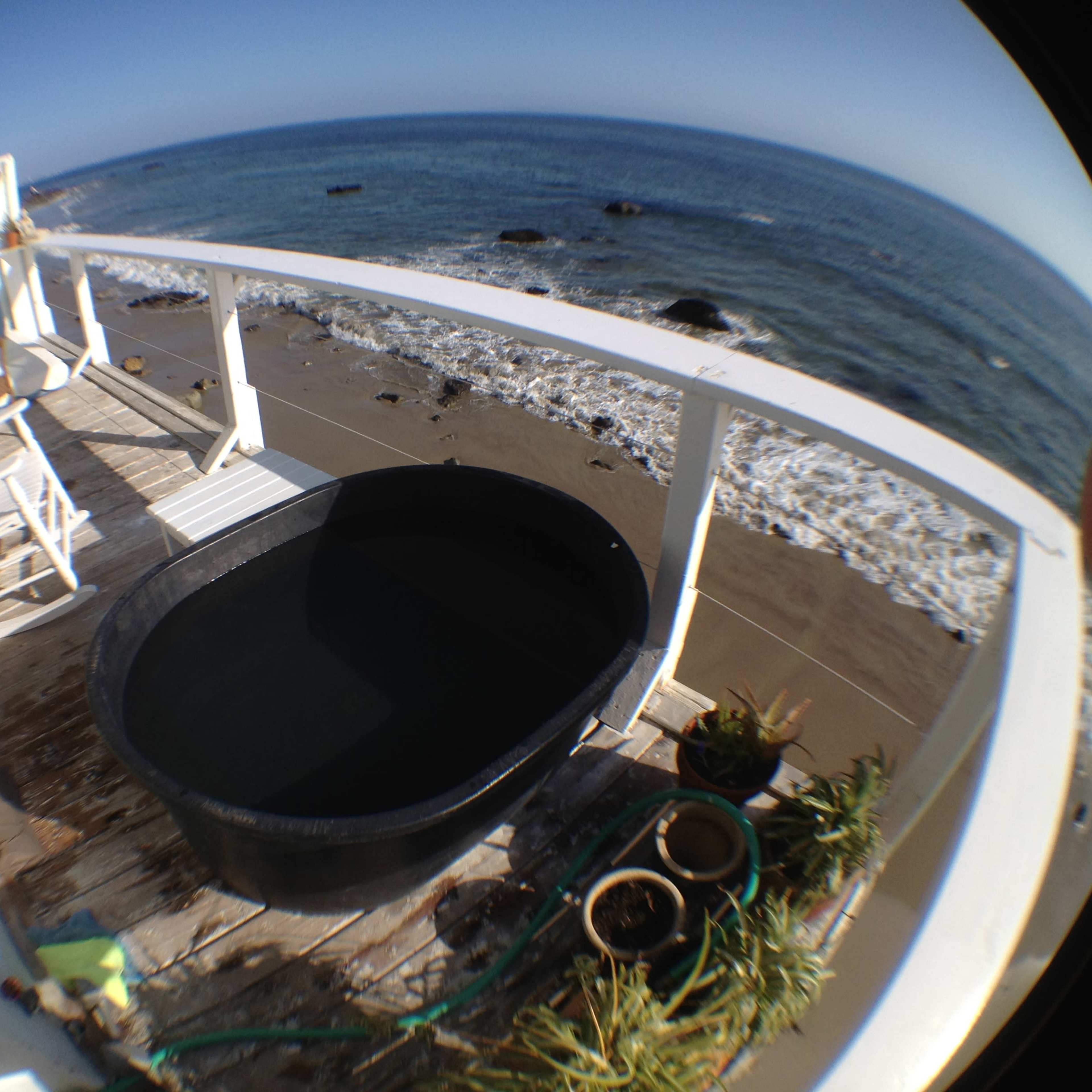 A circular black tub sits on a wooden deck overlooking a sandy beach and ocean waves.