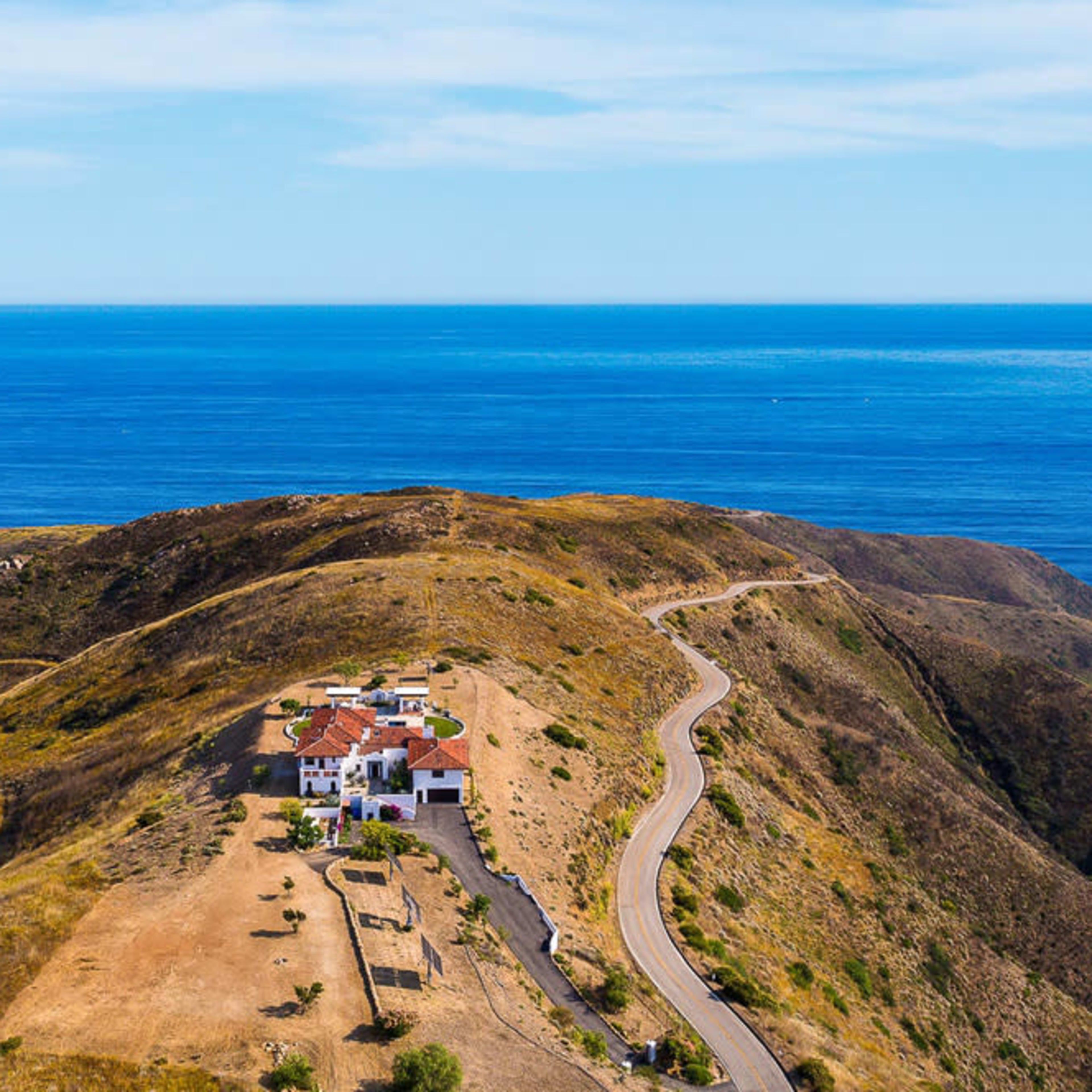 A coastal hillside features a white house surrounded by dry terrain and a winding road leading down toward the ocean.