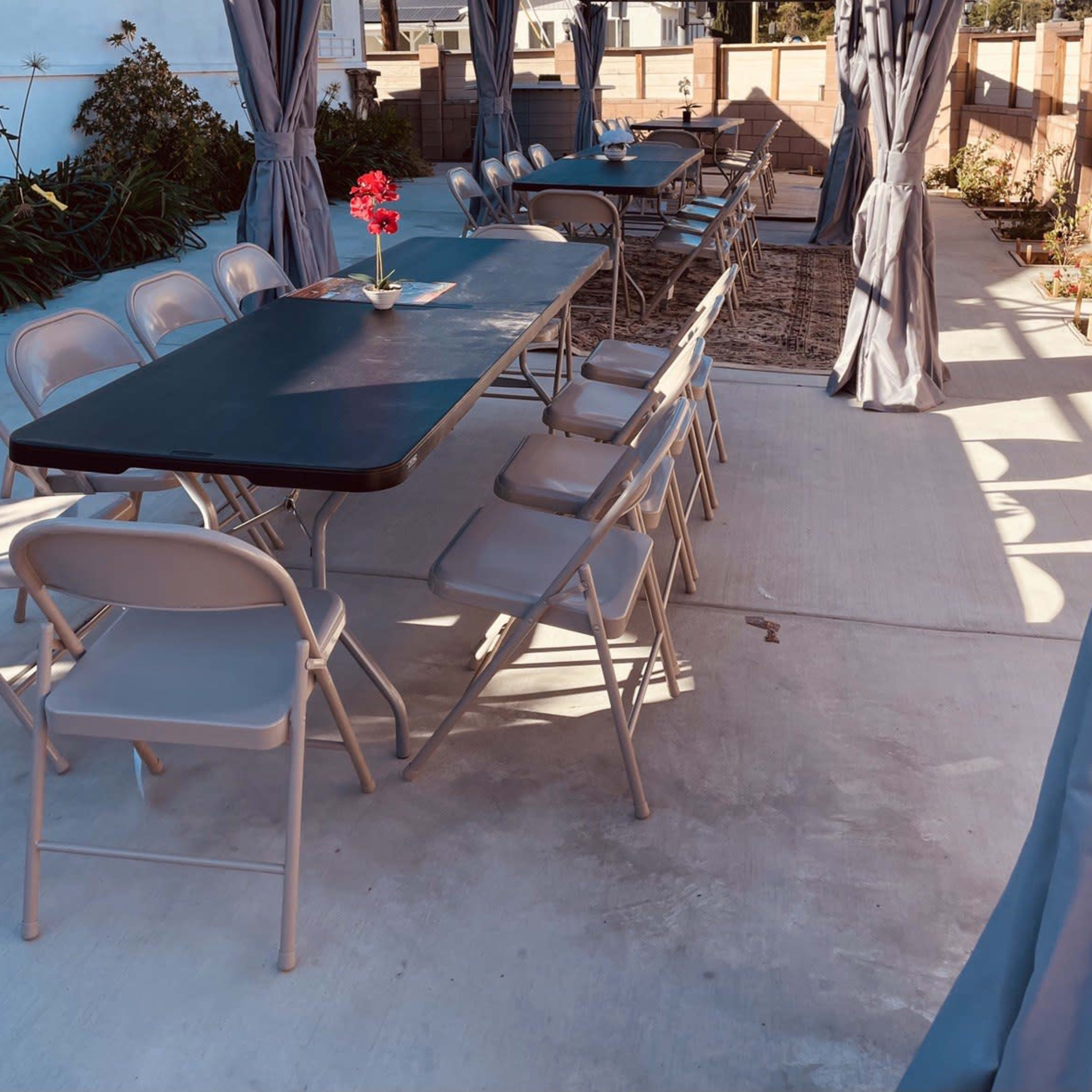 A covered outdoor seating area features several metal tables and folding chairs arranged on a concrete surface, with a single potted flower on one table.