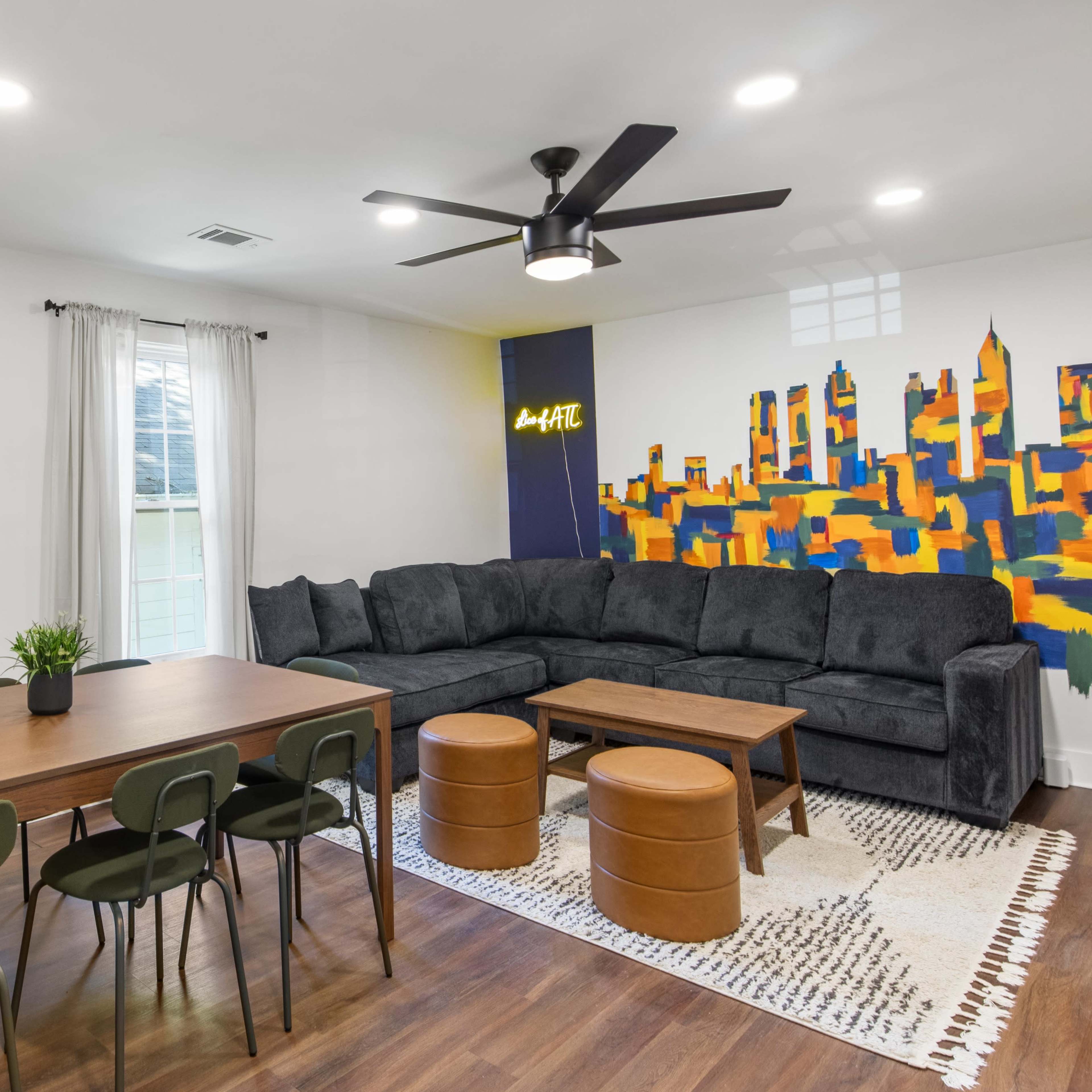The living area features a dark sectional sofa, a wooden dining table with green chairs, and a mural of a city skyline on the wall.
