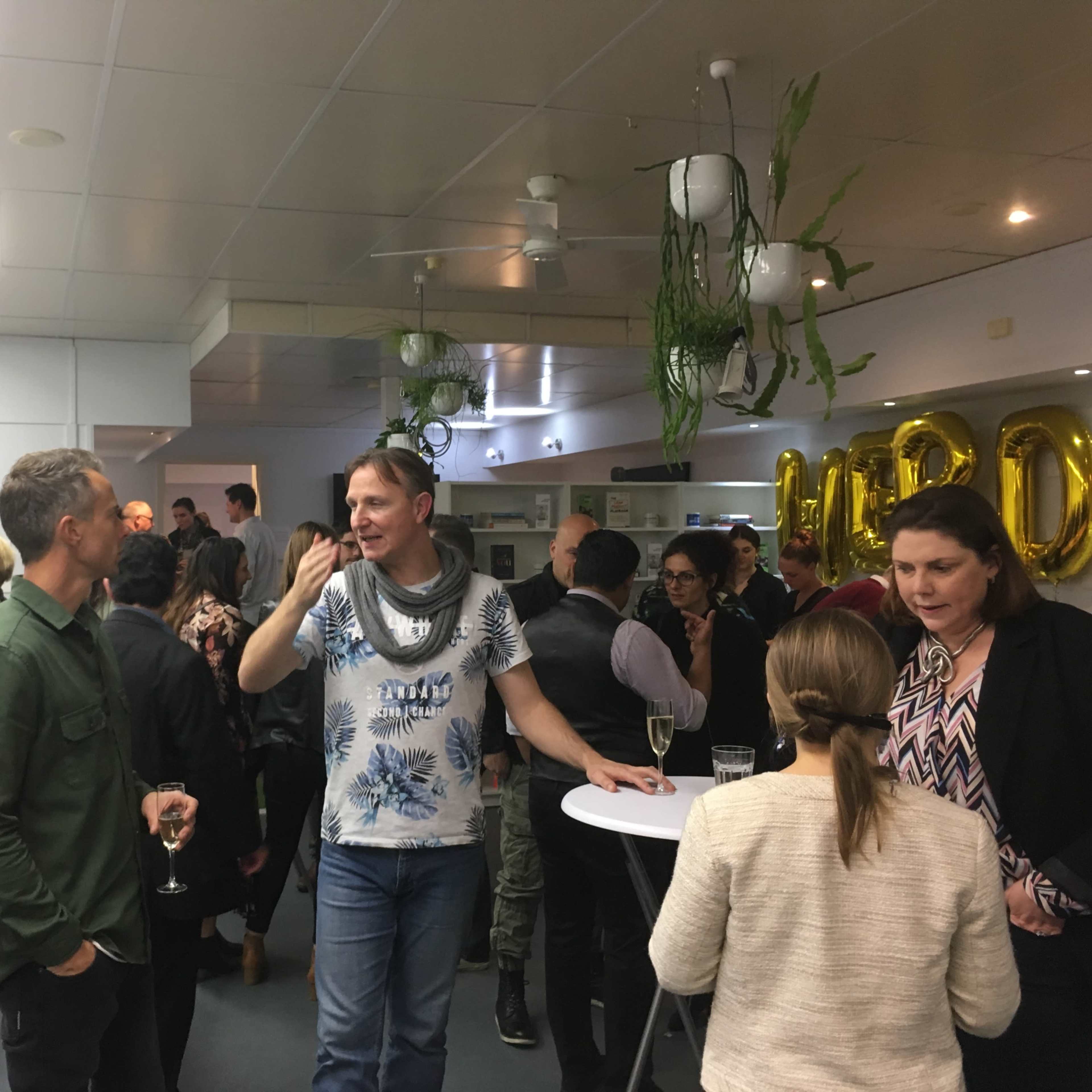 A group of people socialize at a lively event, standing around high tables in a well-lit room adorned with plants.