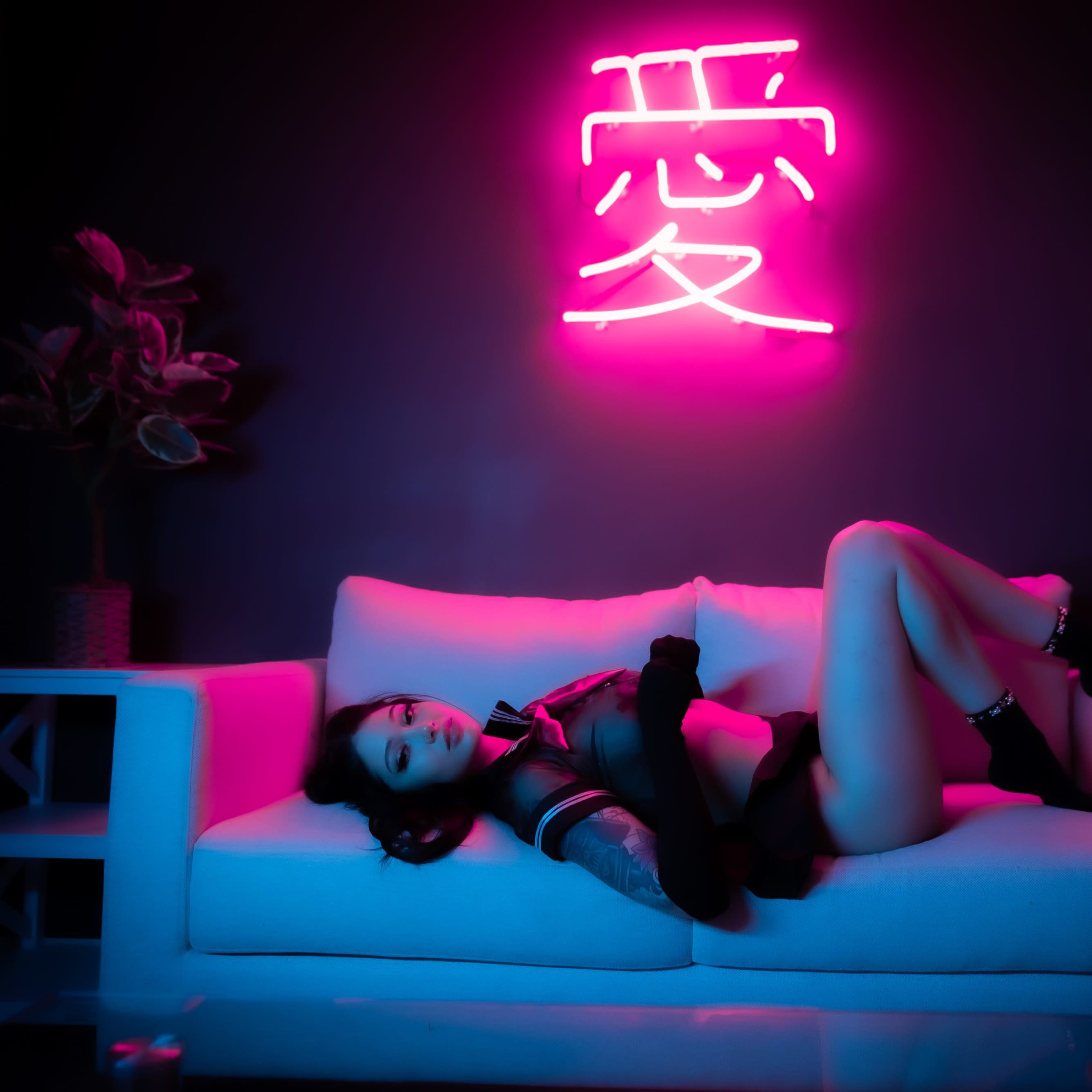 A person lies on a white couch beneath a pink neon sign, with decorative plants and flowers in the background.