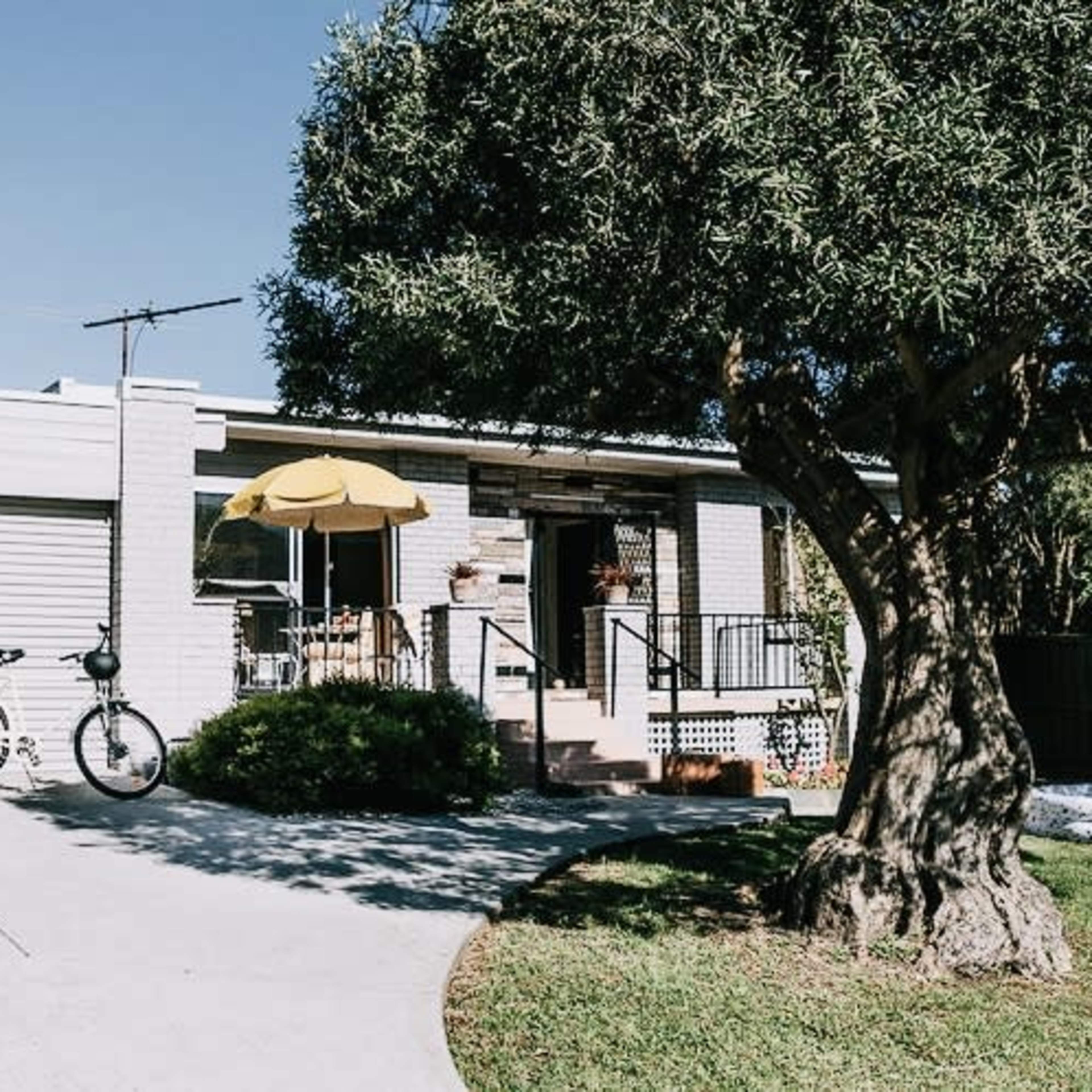 A small, white house with a yellow umbrella over a seating area in the front yard, flanked by a bicycle and greenery.