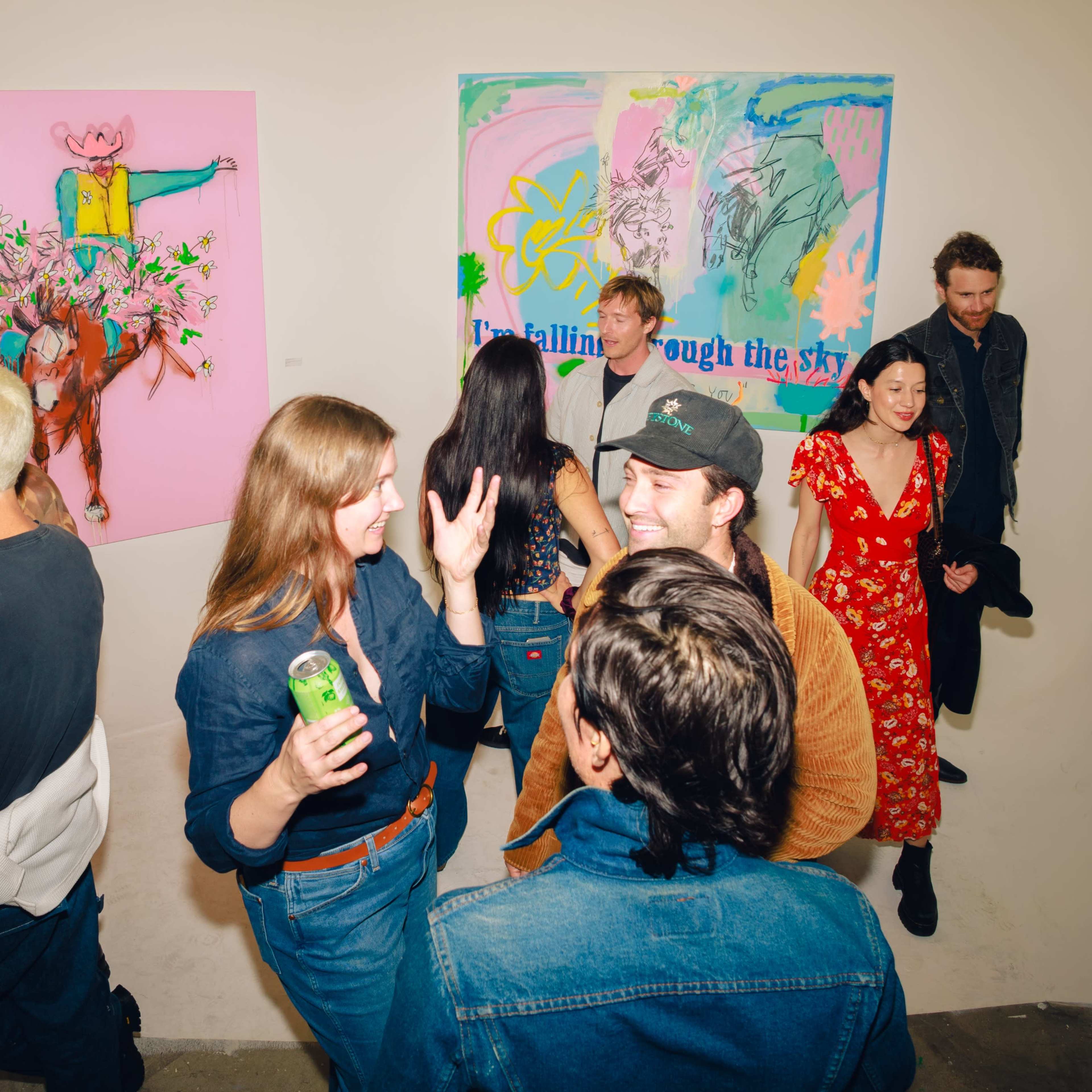 A group of people socialize and enjoy drinks in an art gallery featuring colorful paintings on the walls.