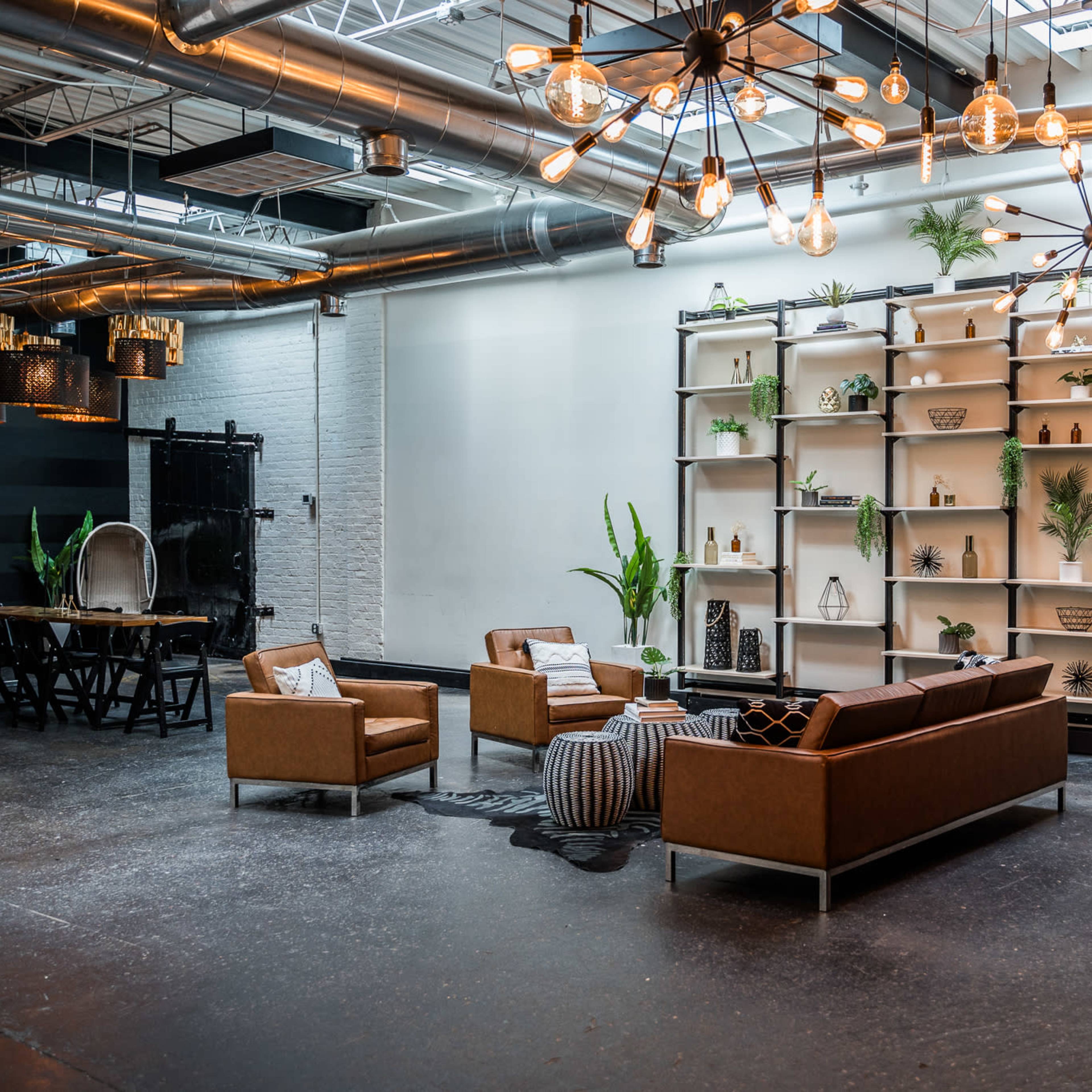 A modern industrial-style interior with a seating area featuring a brown leather couch, two armchairs, and a coffee table, alongside a decorative shelf and a dining table in the background.