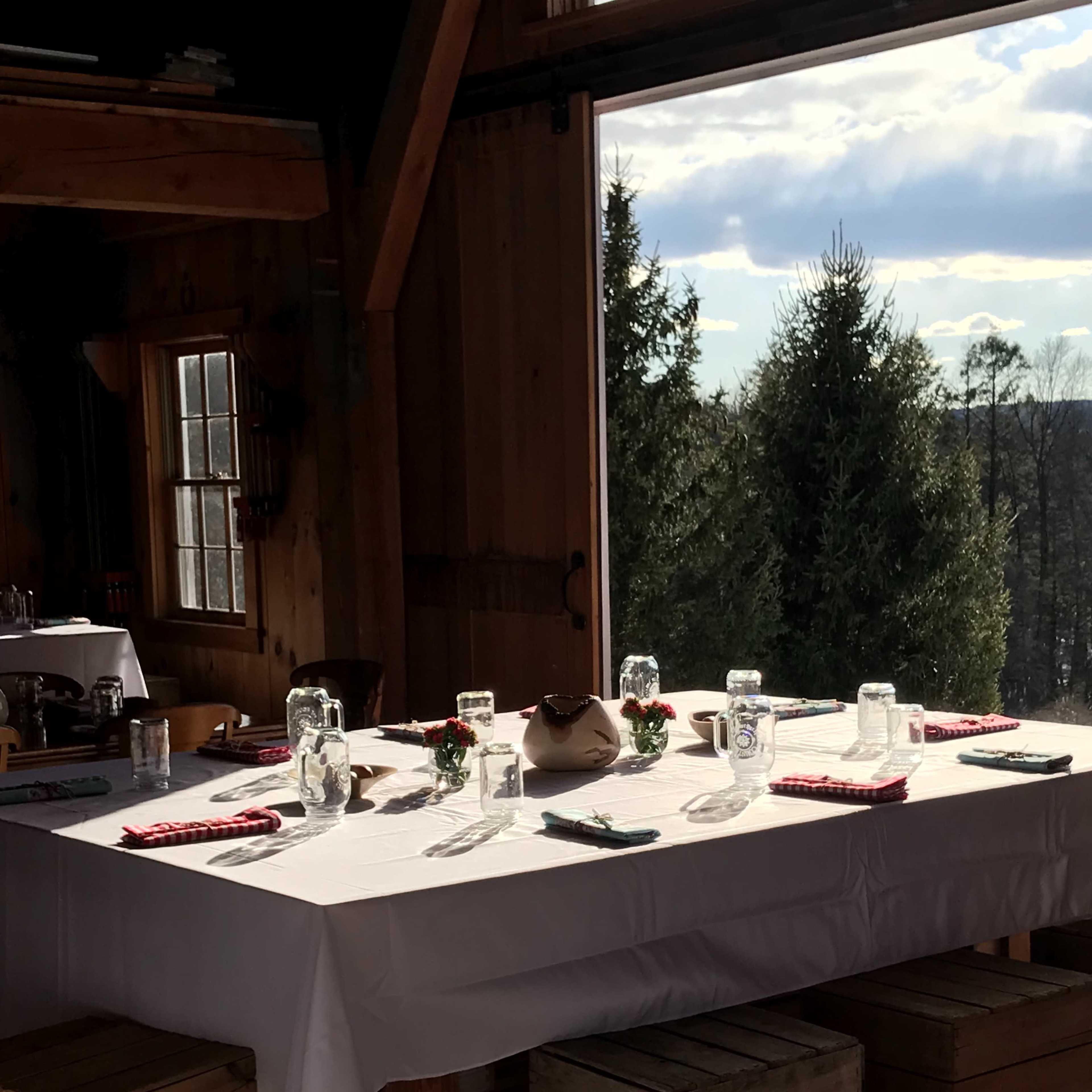 A dining table is set with glassware and utensils inside a wooden cabin, overlooking a forested landscape through large windows.
