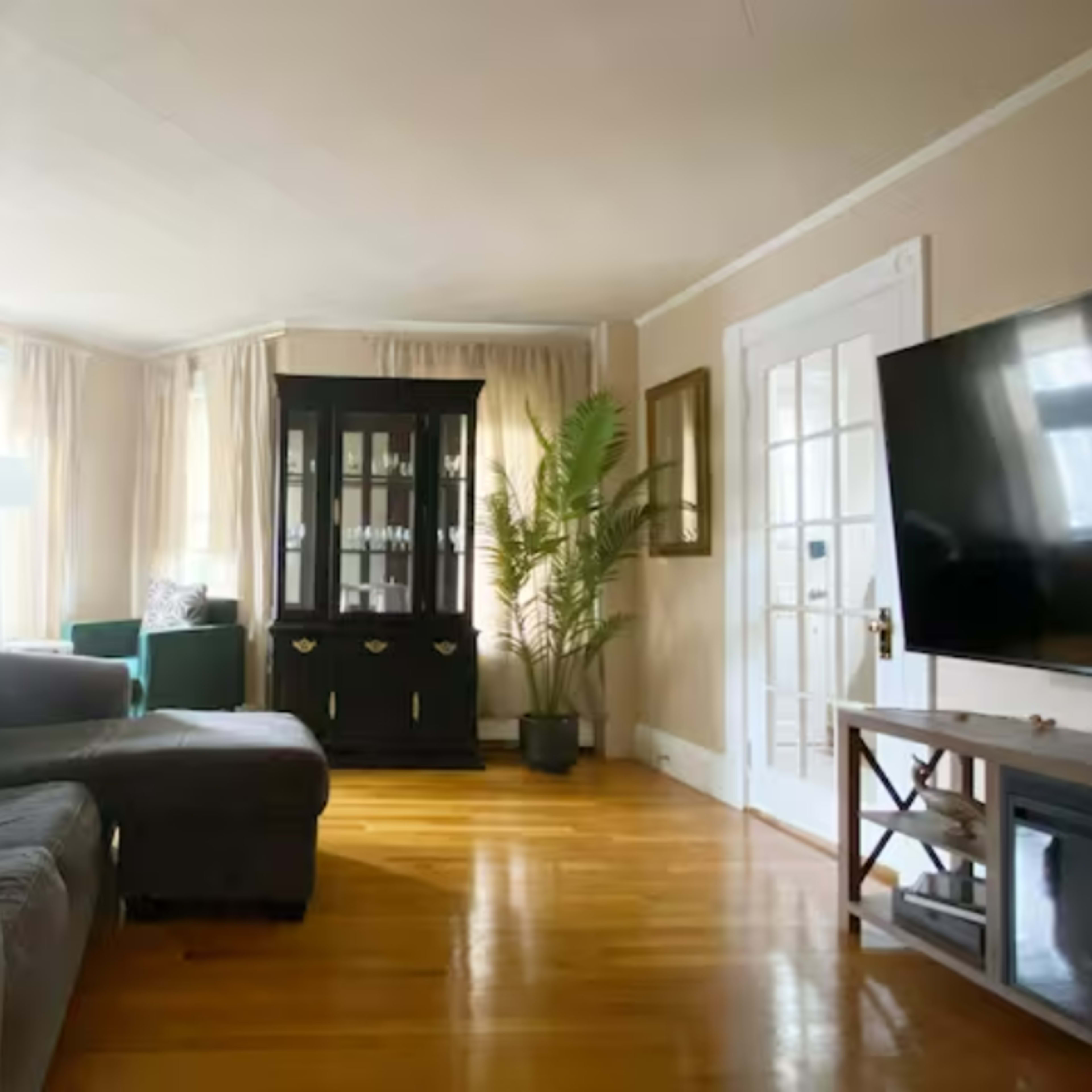 A cozy living room featuring a dark sofa, a small white side table, a black cabinet, and a television on a wooden stand, with large windows dressed in light curtains.