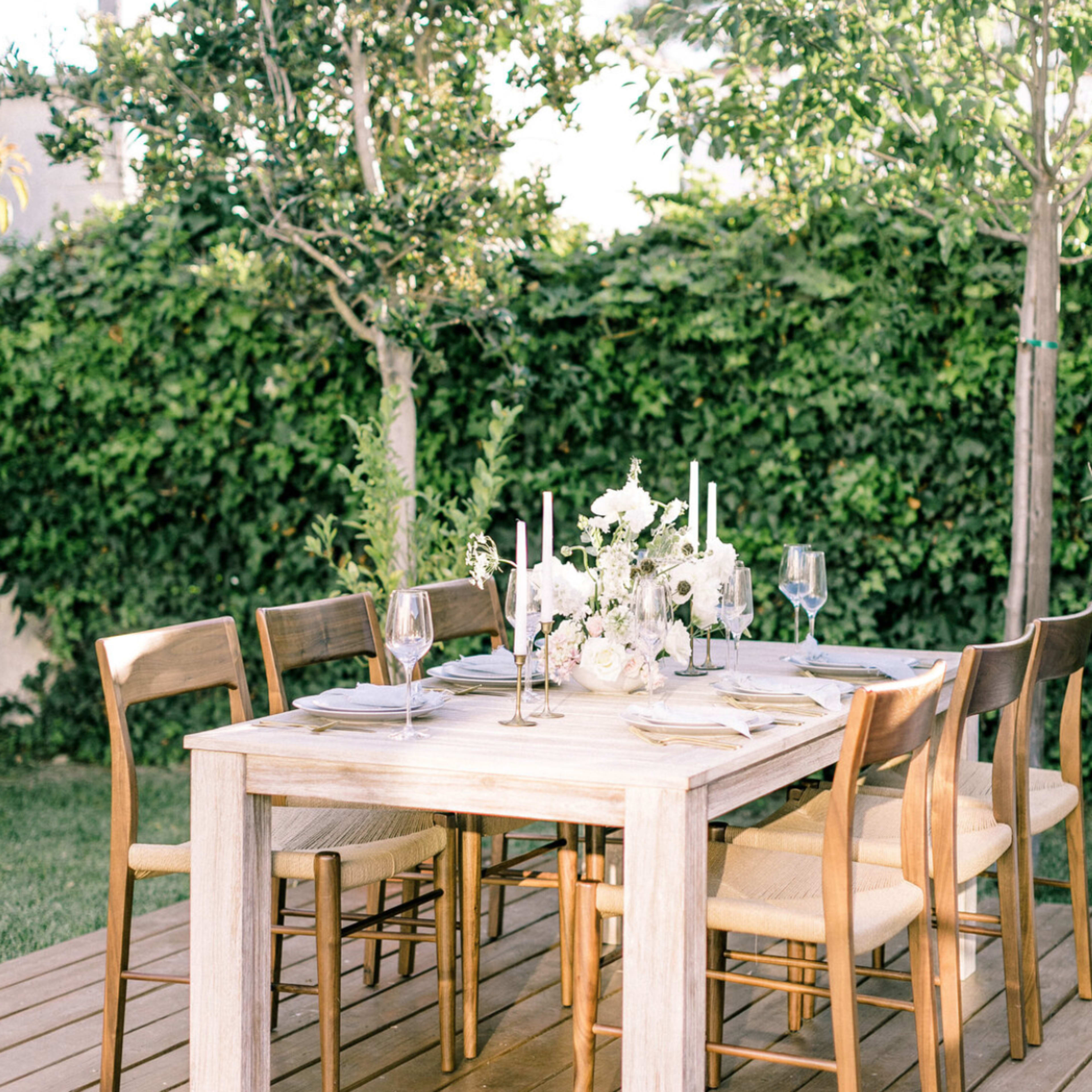 A wooden dining table is set outdoors, surrounded by chairs, with a floral centerpiece and candle holders atop a white tablecloth.