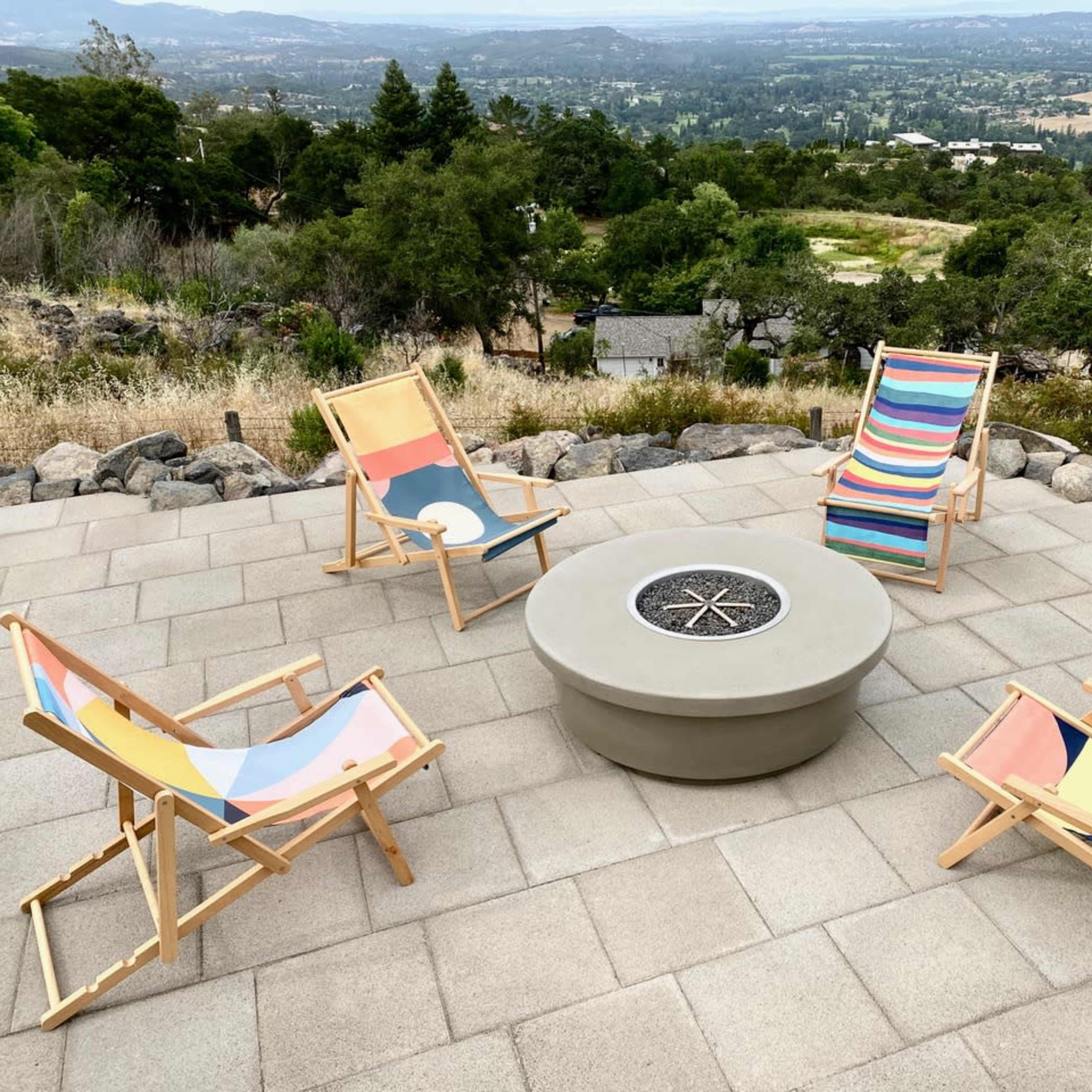 Four deck chairs are arranged around a circular fire pit on a stone patio overlooking a valley.
