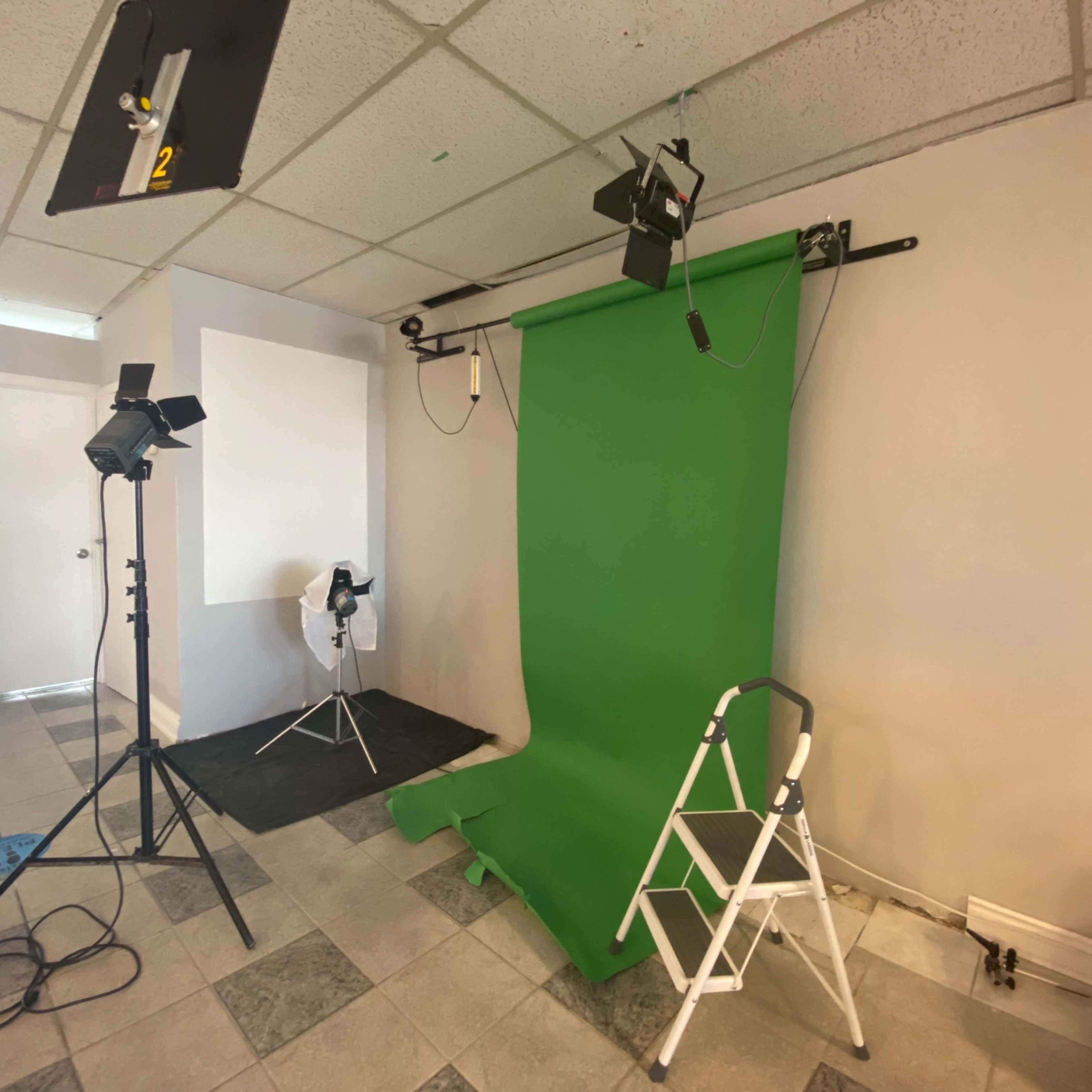 A photography studio featuring a green screen setup, lighting equipment, and a step stool against a plain wall.