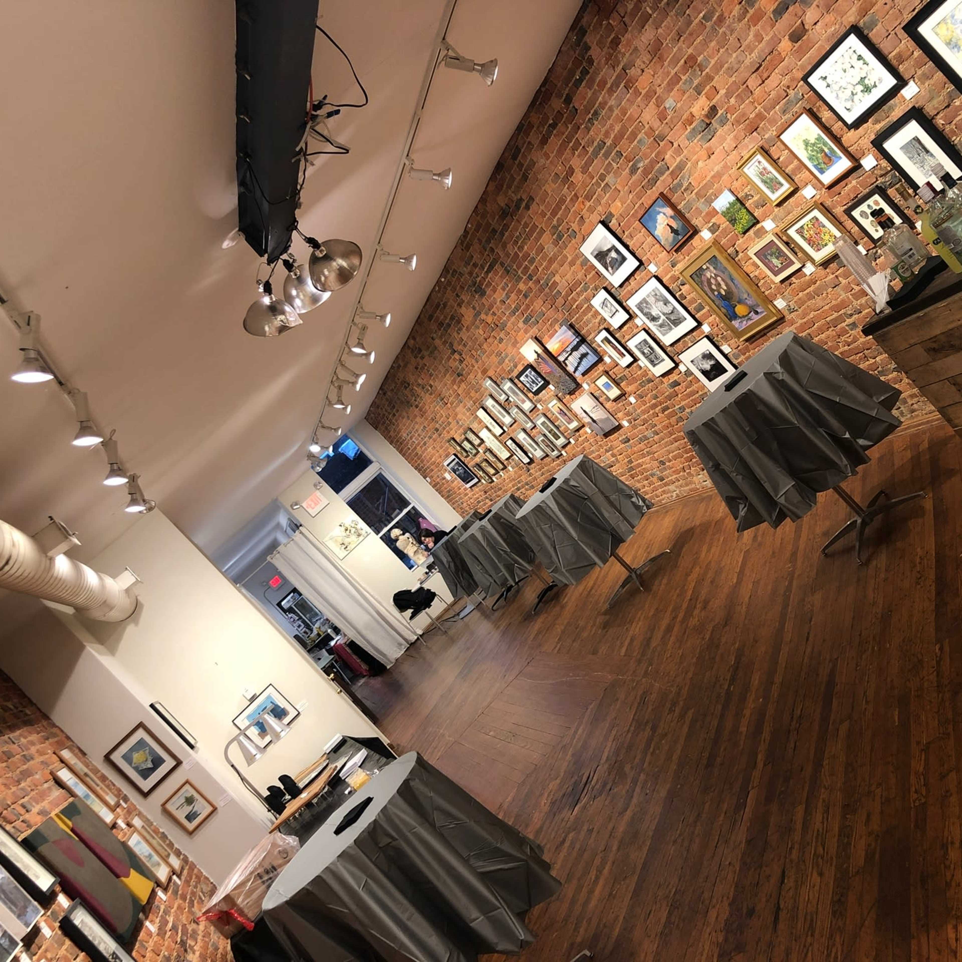 A gallery space features exposed brick walls adorned with framed artworks, and several covered tables are positioned throughout the wooden floor.