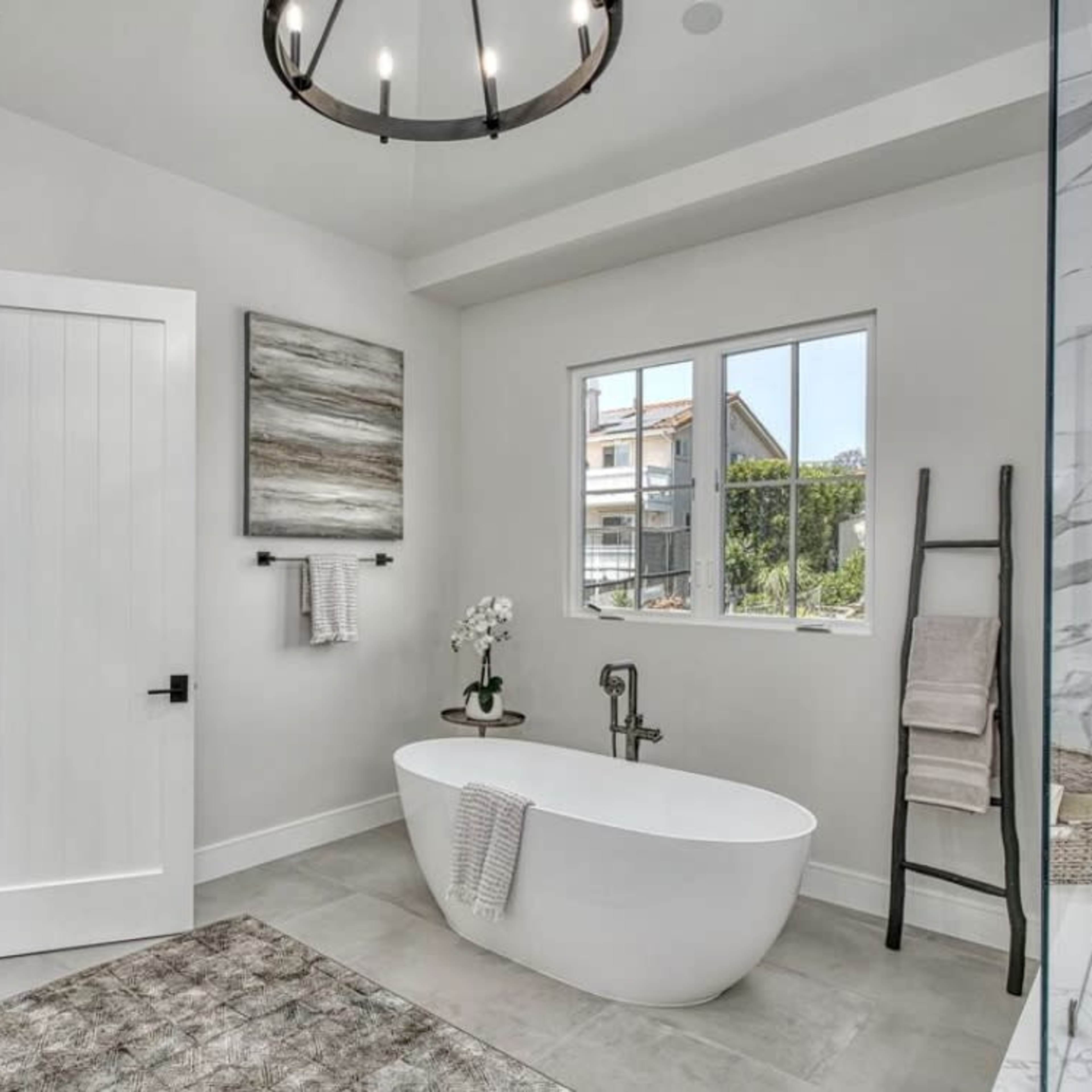 A modern bathroom features a freestanding bathtub, a large window, and a door leading to a bedroom, complemented by neutral colors and contemporary decor.