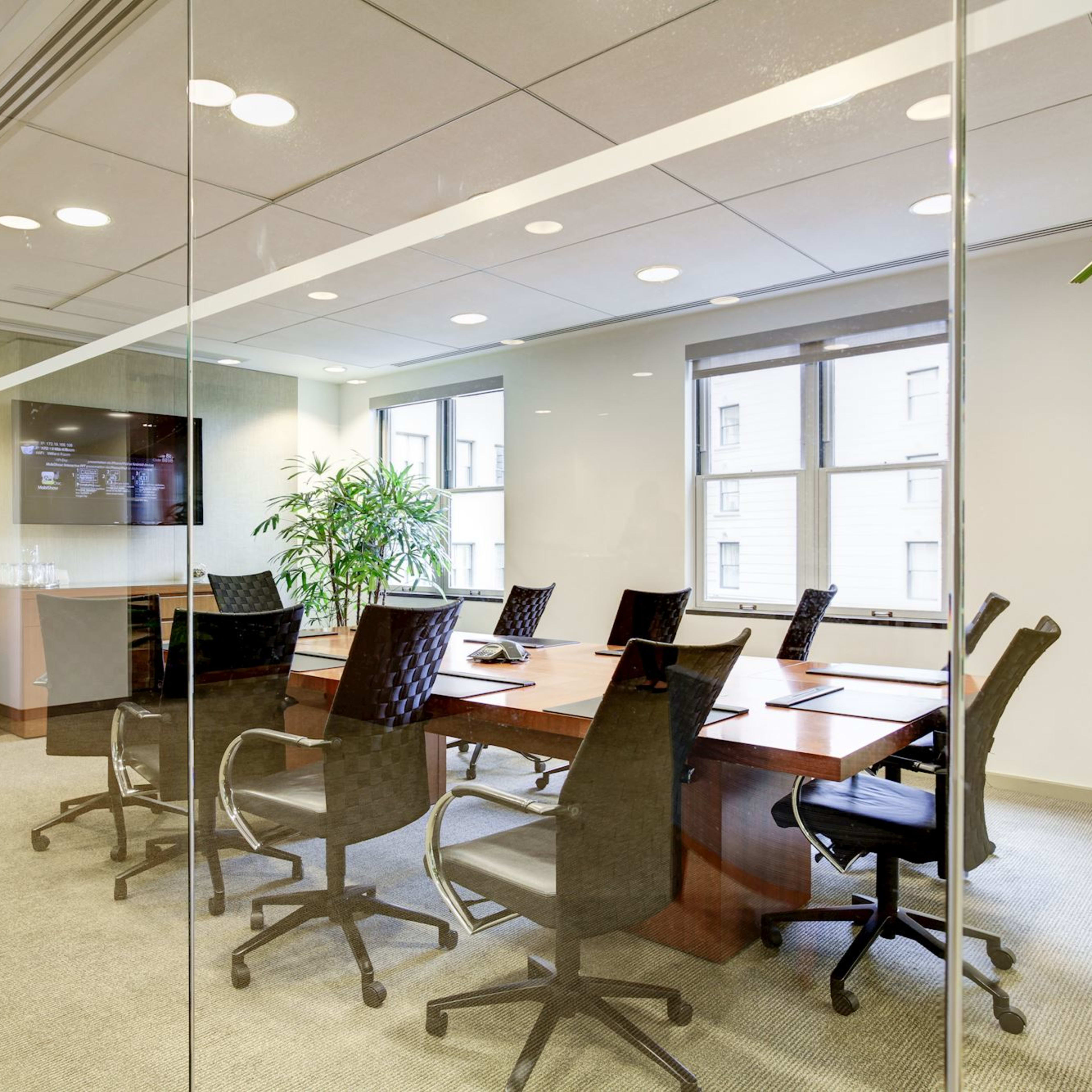 A modern conference room with a large table surrounded by rolling chairs is separated from a hallway by glass walls, featuring a plant and a wall-mounted screen.