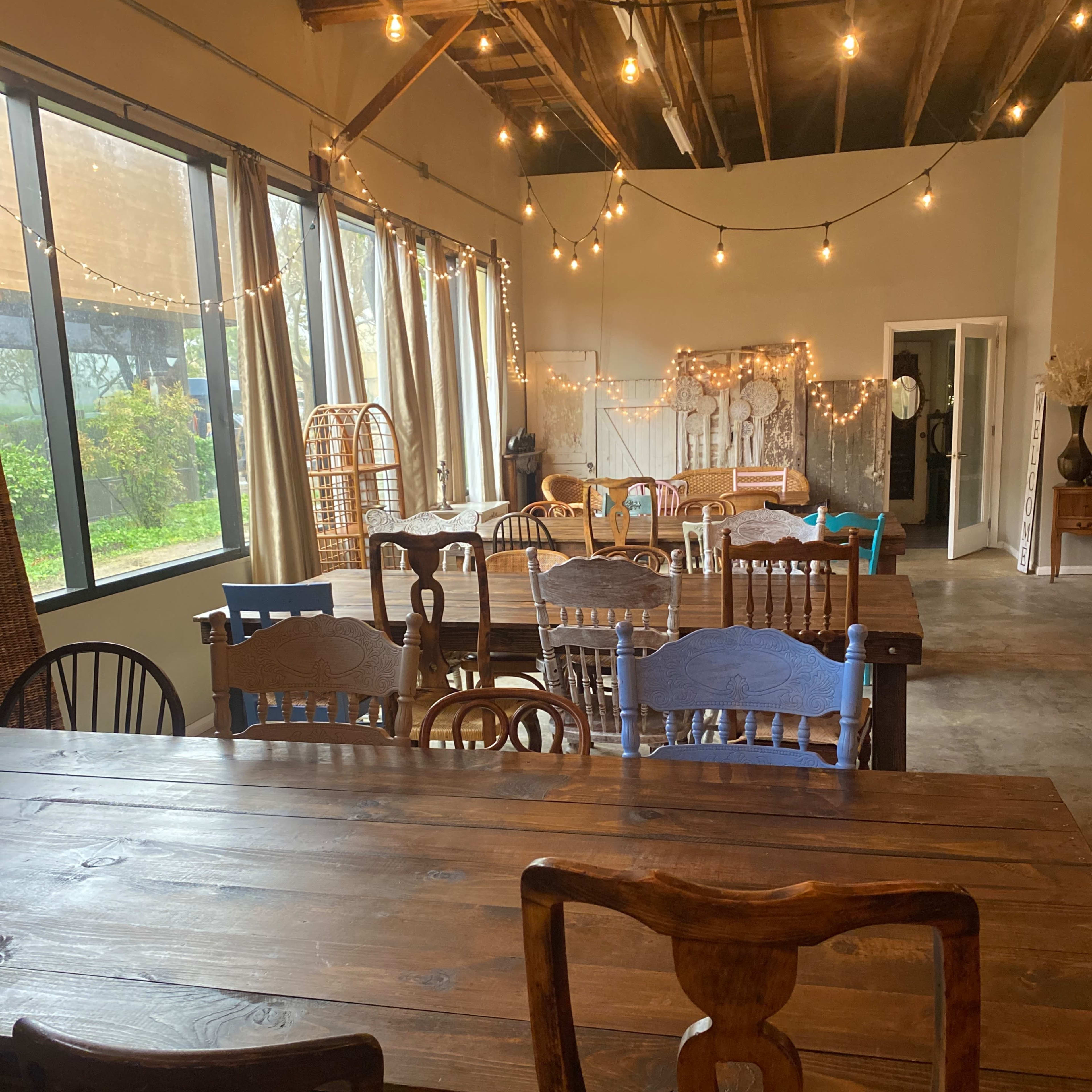 A spacious interior room with wooden tables and chairs, large windows, and string lights overhead.