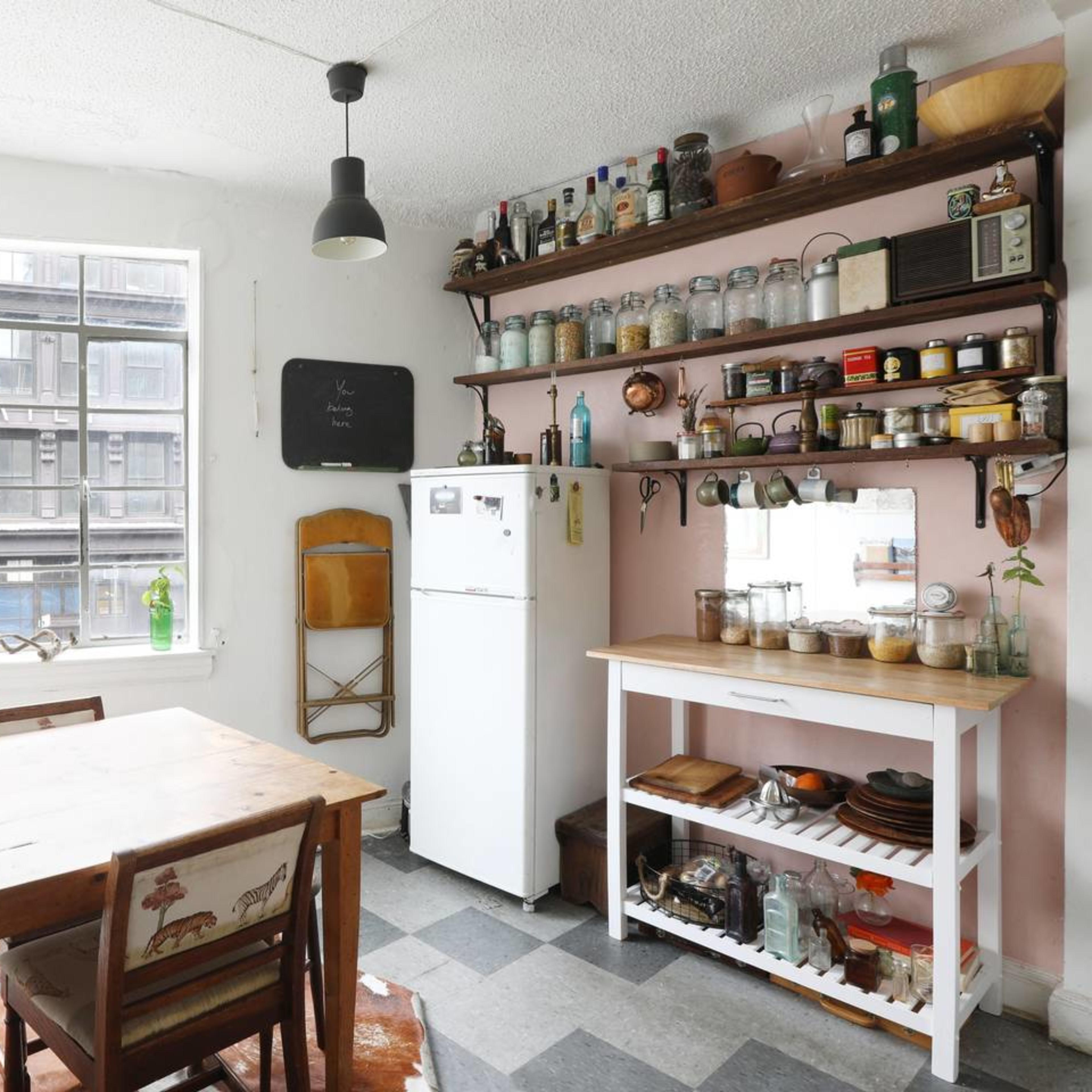 A bright kitchen features a wooden dining table, a white refrigerator, and shelves filled with jars and kitchenware, all under industrial-style pendant lights.